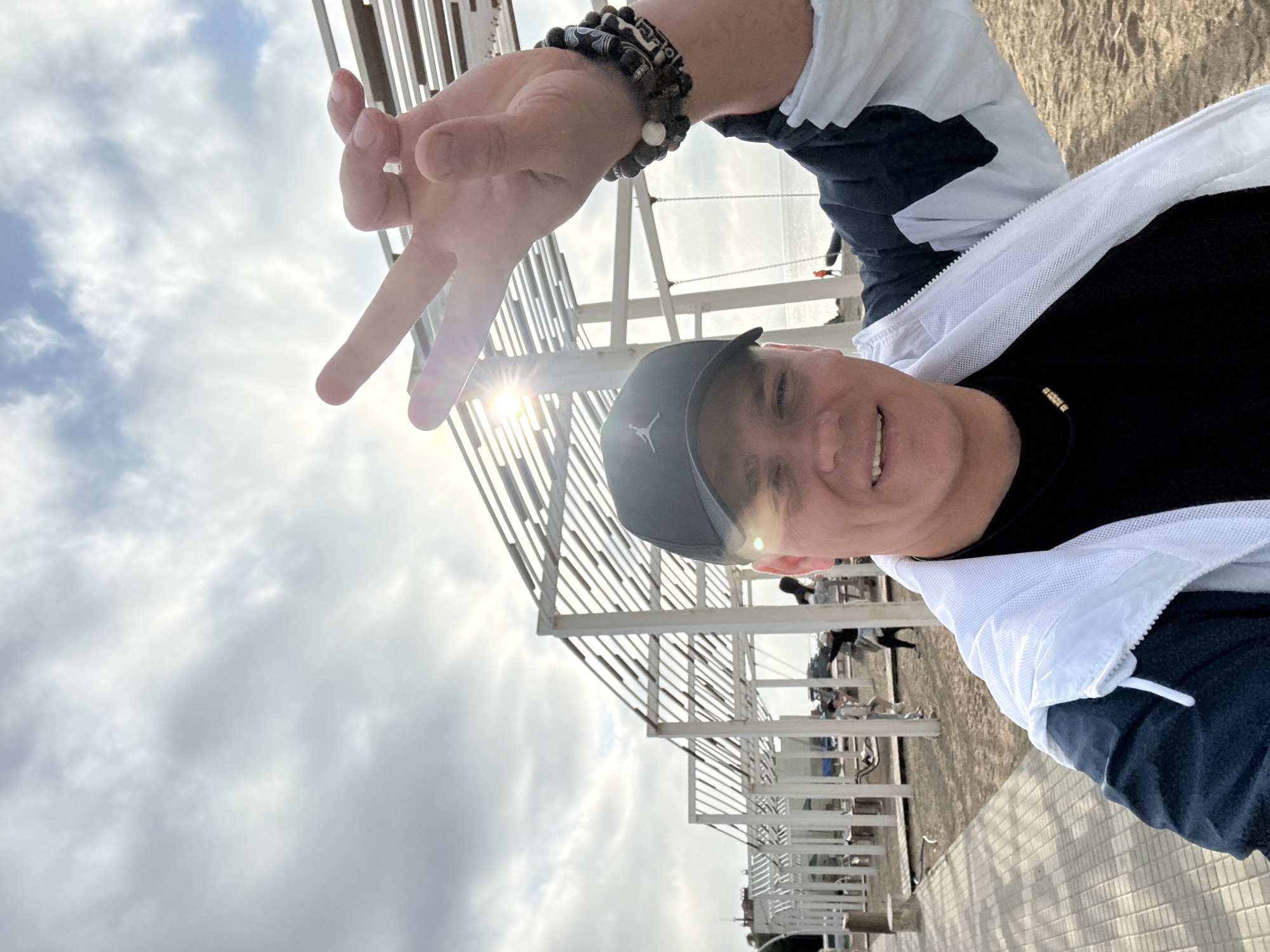 A man wearing a black baseball cap, black t-shirt, and a white and blue jacket is taking a selfie outdoors. He is smiling and holding up his left hand in a peace sign. The background shows a beach with white wooden structures, possibly beach shelters or pergolas, and the ocean. The sky is overcast with some patches of blue. The man is also wearing several beaded bracelets on his left wrist. The overall lighting suggests it is daytime. The scene appears to be a recreational area on a beach.