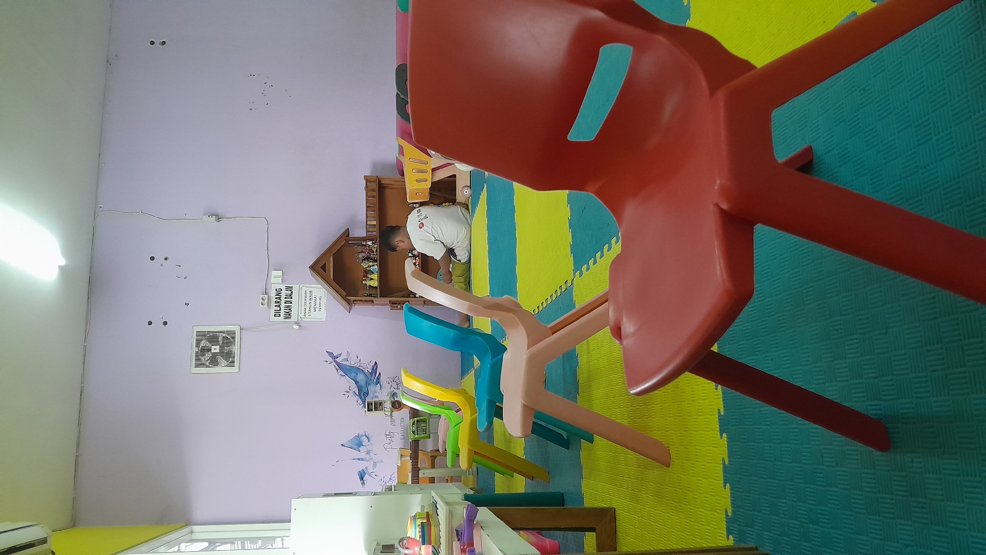 The image depicts the interior of what appears to be a classroom or play area in Kasiman, Indonesia. In the foreground, a bright red plastic chair is prominently featured, with its legs resting on a blue and green interlocking puzzle mat. Behind it, a row of four smaller plastic chairs, colored orange, yellow, blue, and light pink, are lined up on the same matting.
Further in the background, a young boy, appearing to be of school age, is seated on the floor, facing away from the camera. He is wearing a white t-shirt with some text or graphic on the back and is engaged with toys on a wooden shelf unit behind him. This shelf unit holds various small figurines and toys. To the left of the boy, a wooden dollhouse structure is visible.
The walls are painted a light purple hue. On the wall, there is a white ventilation fan, and a sign with text in a foreign language, appearing to be a prohibition against eating inside. There are also several small markings and holes on the wall, possibly from posters or decorations. Adjacent to the chairs, on the left side of the image, a white table surface with colorful toys and containers is partially visible. The lighting suggests an indoor environment, with a ceiling light fixture visible at the top of the frame. The overall atmosphere is that of a children's educational or recreational space.