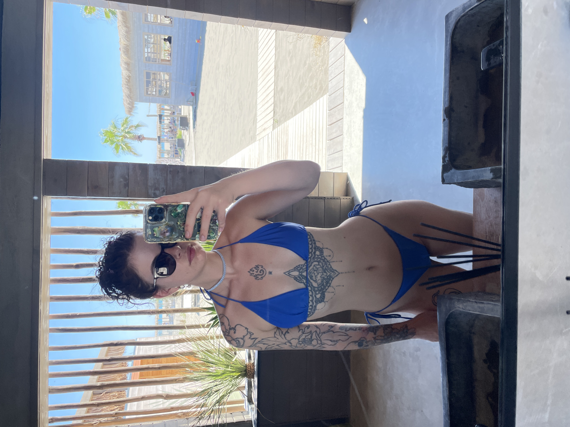 A young woman, viewed from the chest down, stands in front of a mirror at what appears to be a beachside establishment in Donji Štoj, Montenegro. She is wearing a blue bikini and sunglasses. Tattoos are visible on her arm and torso, including intricate designs on her abdomen. She is holding a mobile phone in her right hand, taking a selfie. The background, reflected in the mirror, shows a bright, sunny day with a sandy beach, palm trees, and a light-colored building with a thatched roof. Wooden walkways are visible on the sand. The foreground of the image shows the top of a dark sink or basin with several dark sticks standing upright within it.