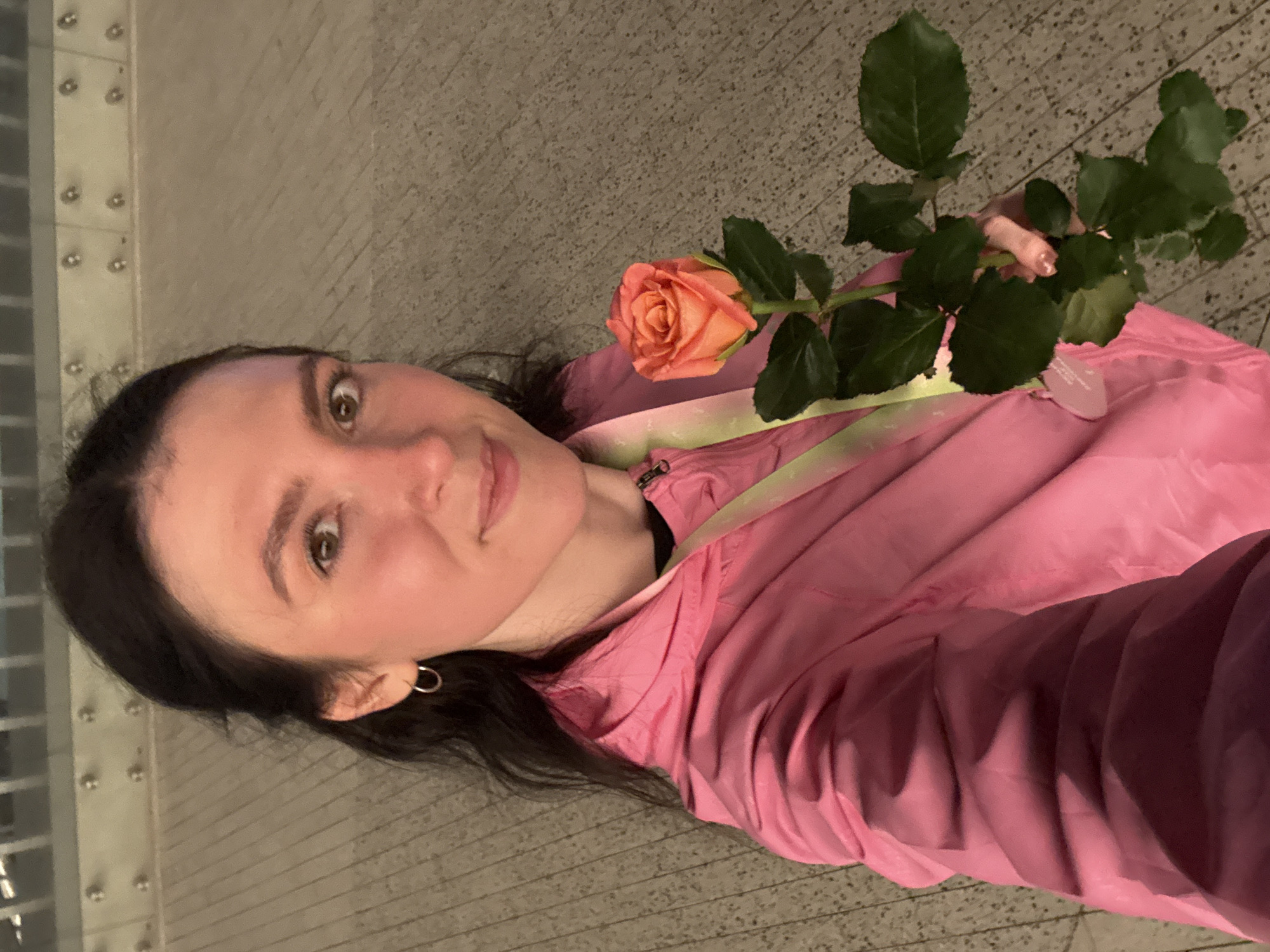 A woman is posing in a selfie. She holds a single peach rose. She wears a pink jacket with green accents. She has dark hair and wears a silver hoop earring. The background is a grey concrete surface with a textured pattern. A metallic structure with evenly spaced bolts can be seen to the left. The location cannot be definitively identified as Kazan, Russia.