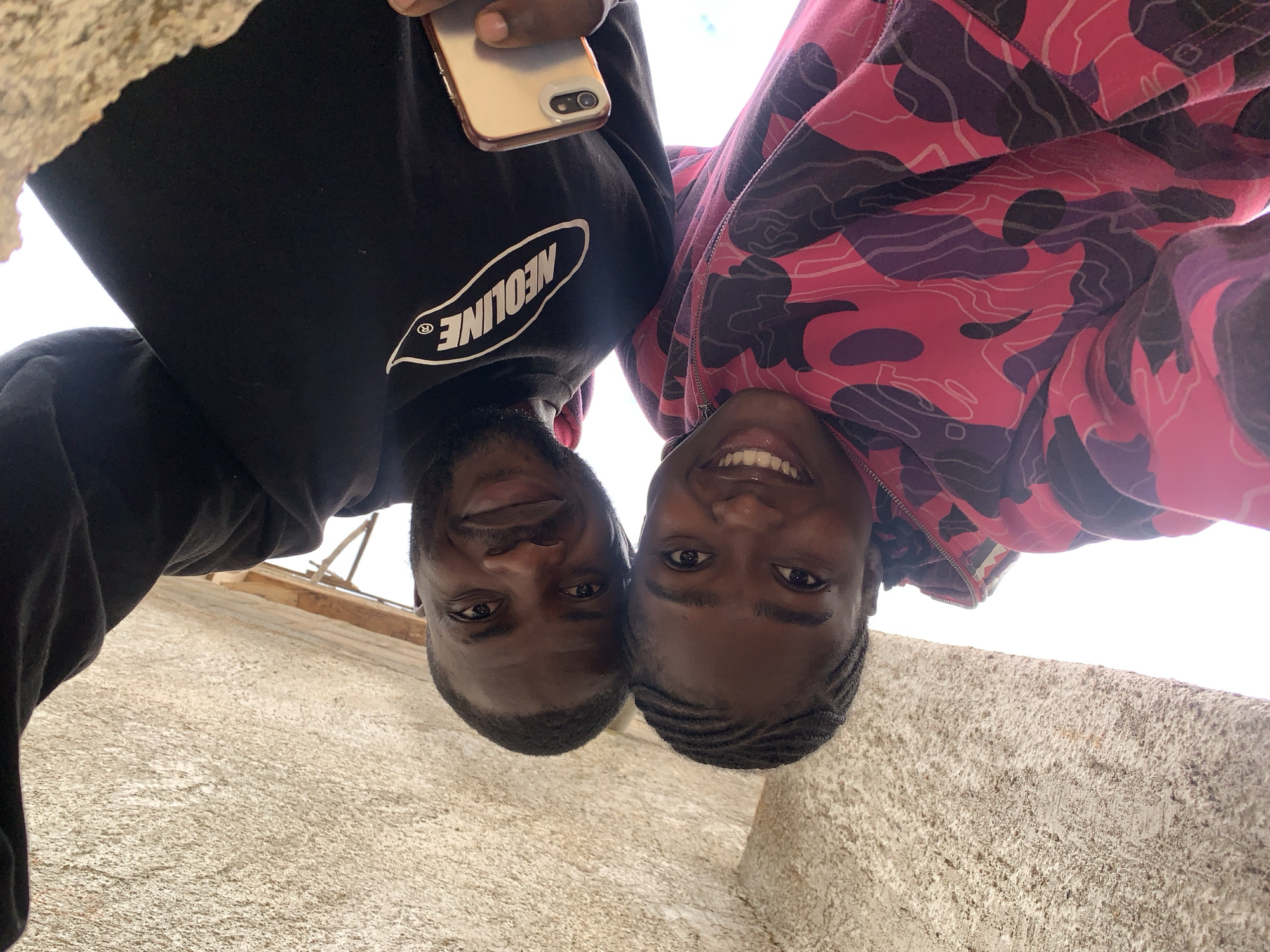 Two individuals are depicted from a low-angle perspective, looking directly down towards the camera. The scene takes place in Rafin Par, Nigeria. The individual on the left is a male with dark skin and a short beard, wearing a black long-sleeved shirt featuring a white oval logo with the word "NEOLINE" in capital letters. He holds a light-colored smartphone with a dual-camera lens visible in his right hand. His expression is a slight smile. The individual on the right is a female with dark skin and braided hair, wearing a hooded jacket with a pink, dark purple, and black camouflage pattern. She displays a broad smile, revealing her teeth. Their faces are in close proximity at the lower center of the frame. The background consists of a textured, light-beige concrete or stucco wall beneath their faces, and a bright, overcast sky or strong light source above and between their upper bodies. In the upper left portion of the image, faint wooden structures, possibly scaffolding or construction elements, are visible against the bright background.