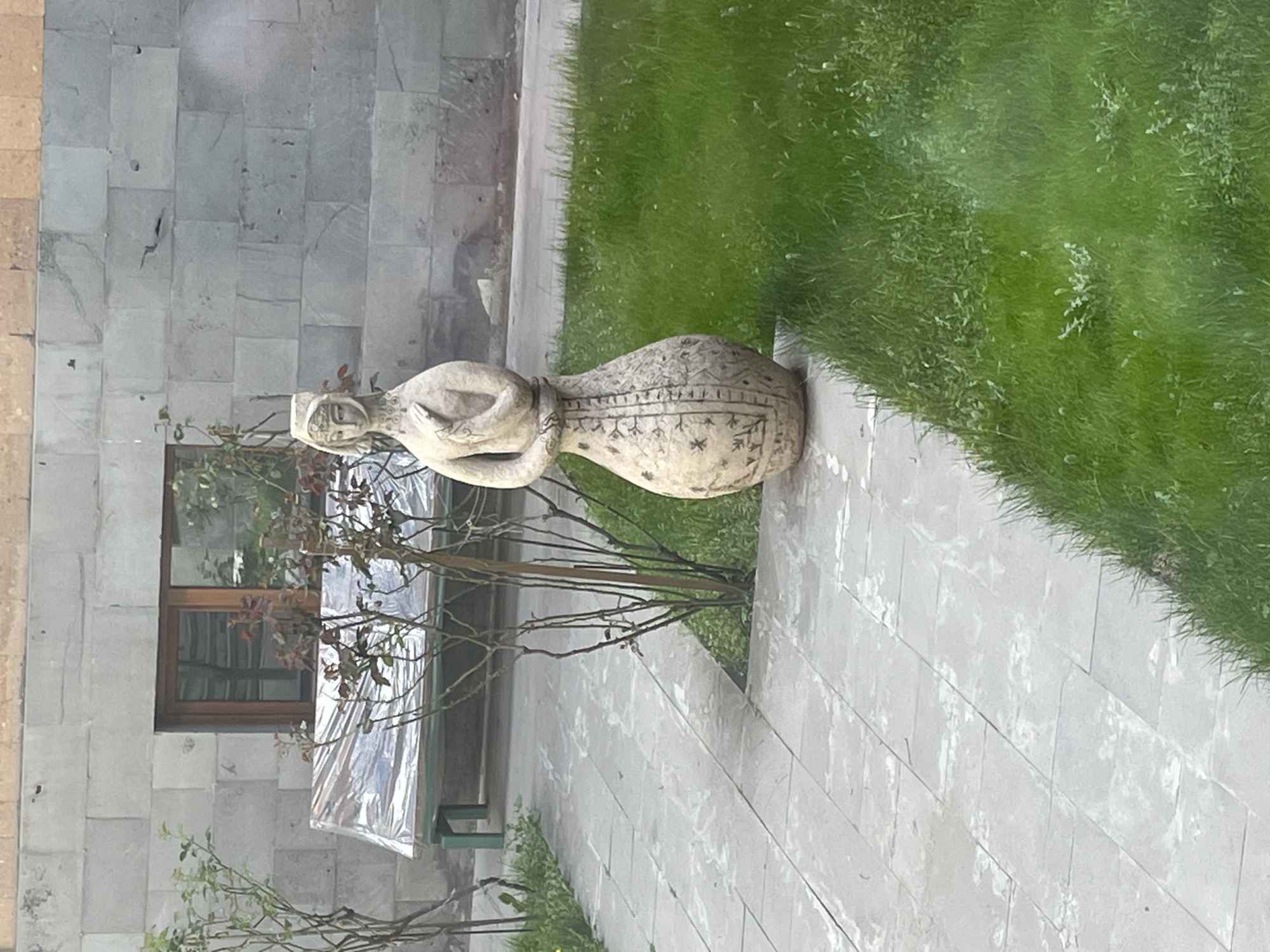 The image shows a light-colored stone sculpture of a female figure, resting on its side on a gray stone-tiled walkway bordering a grassy area. The sculpture has decorative etchings. Behind the sculpture is a windowed building with gray stone walls. A thin tree is also visible. The scene is located in Yerevan, Armenia.