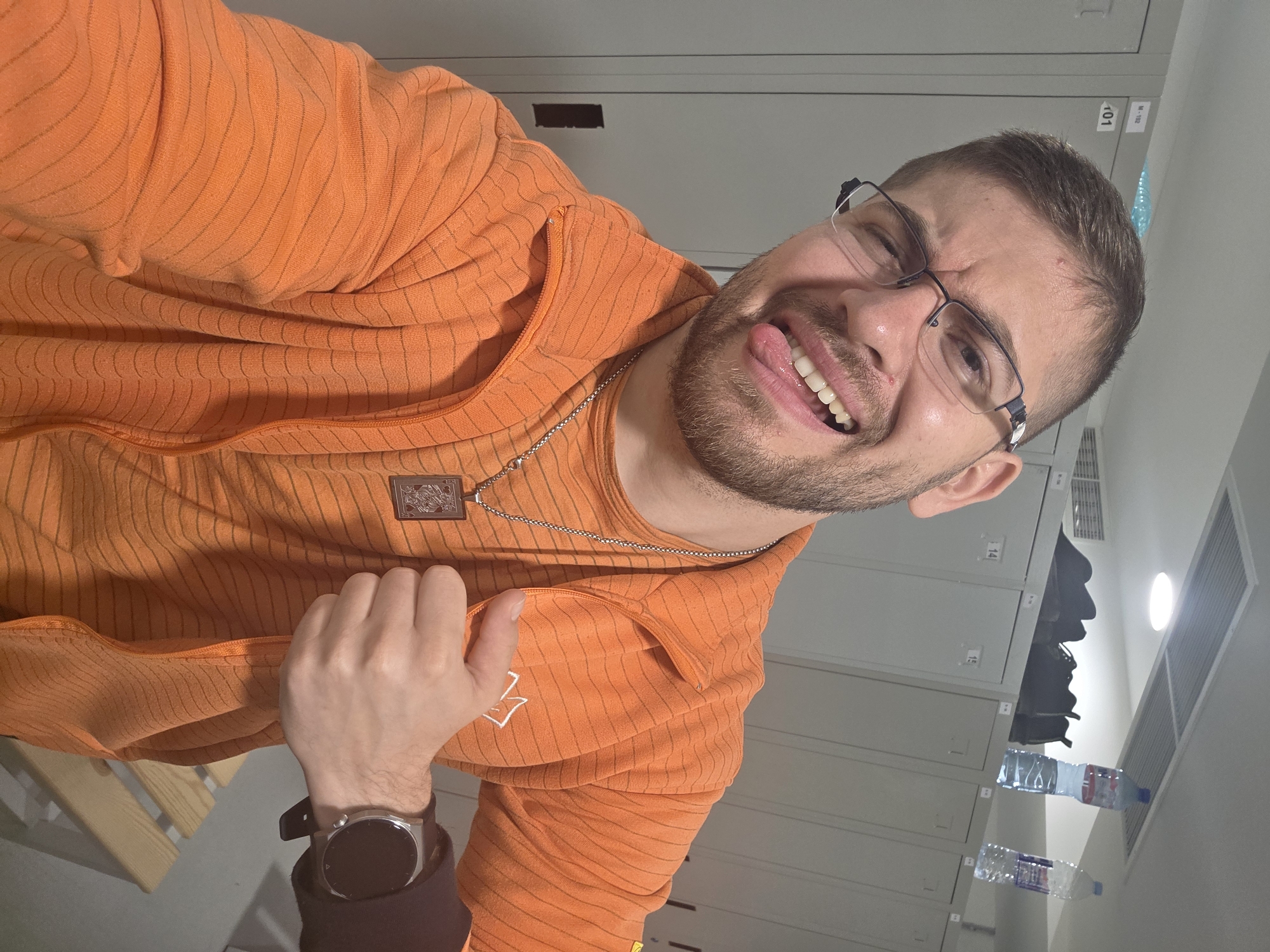 A man is taking a selfie in what appears to be a locker room. He is wearing an orange striped hooded sweater and glasses. He is sticking his tongue out and winking at the camera. He is wearing a smartwatch on his left wrist. The background consists of gray metal lockers. The location cannot be determined as Plovdiv, Bulgaria, based on the image provided.