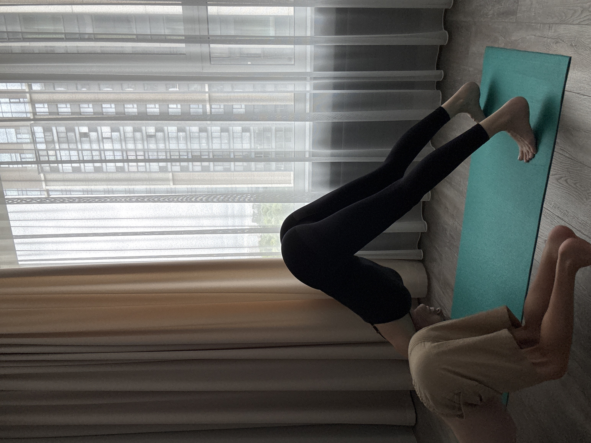 In a room in Khimki, Russia, two individuals are engaged in a yoga session near a window with sheer white curtains, revealing a view of apartment buildings. A turquoise yoga mat is laid out on a wooden floor. One person is positioned in a downward-facing dog pose, wearing black leggings and barefoot. Their body is angled with the buttocks raised. The second individual is partially visible in the foreground, kneeling with their legs bent and resting on the floor, wearing light-colored shorts.