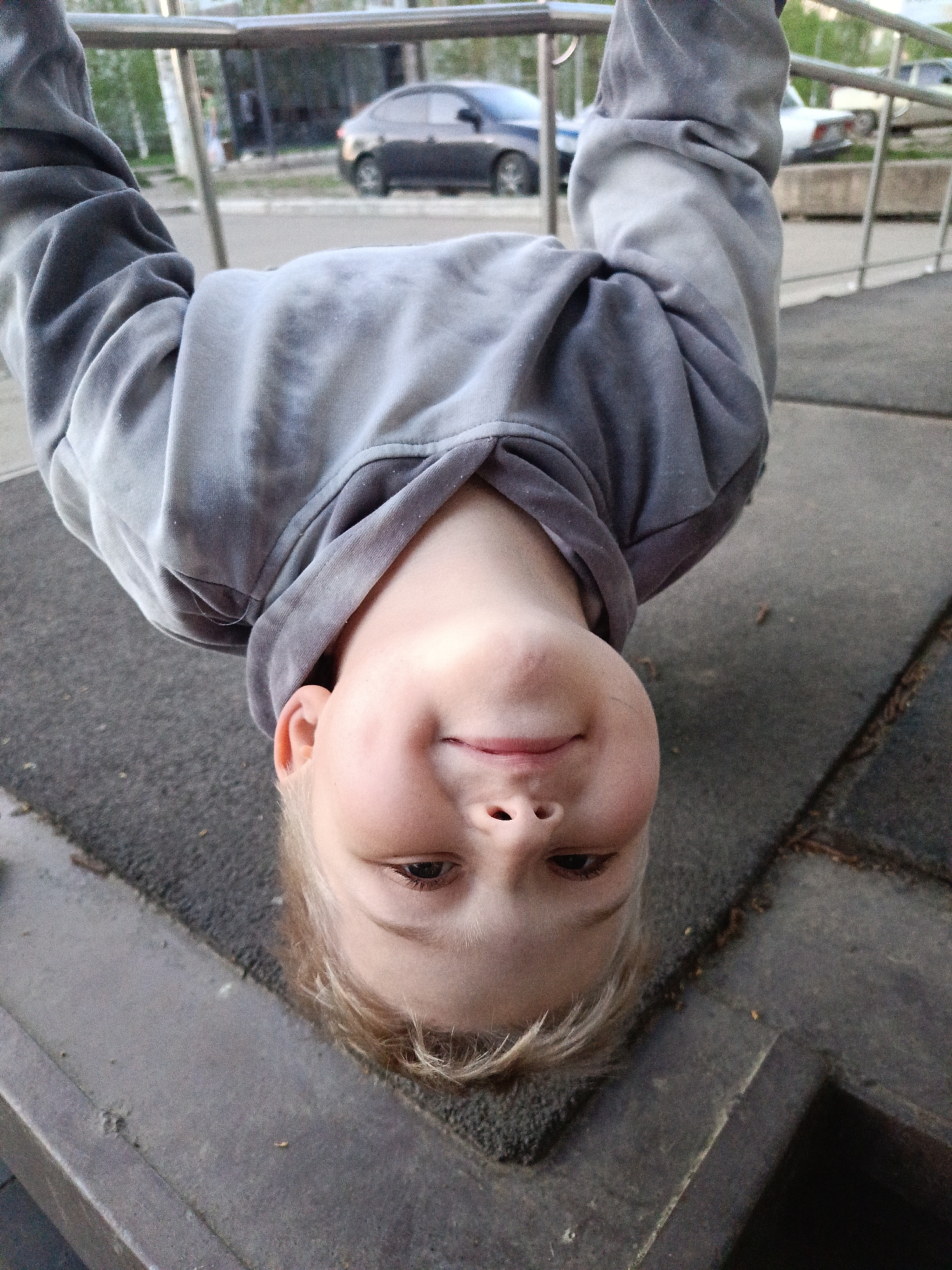 A child is hanging upside down from a metal bar. The child is wearing a grey hooded sweatshirt and has blonde hair. The location appears to be a playground or park, based on the padded ground visible beneath the child. Cars are parked on the street in the background. The city may be Chaikovsky, Russia, as requested.