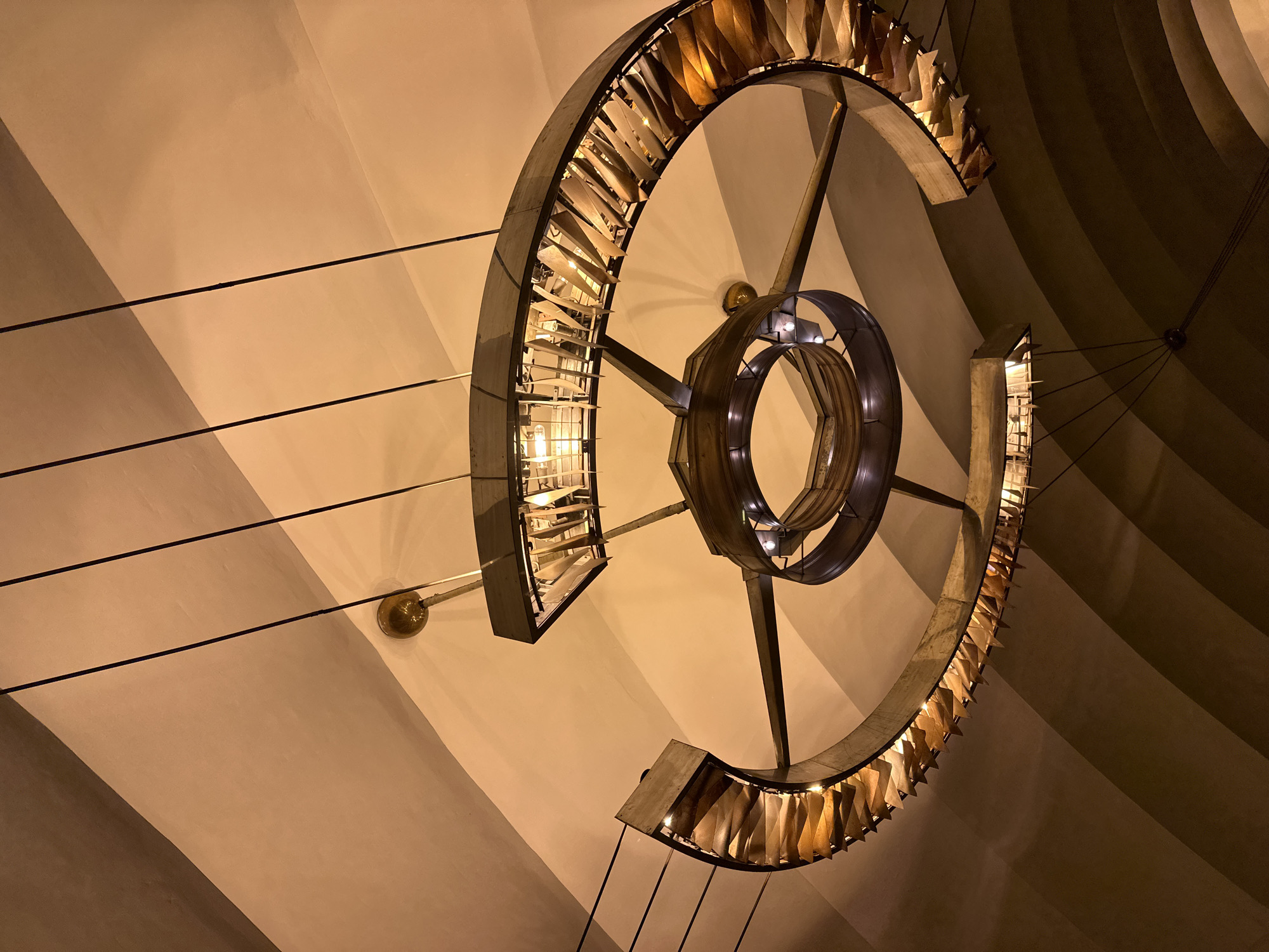 The image captures a low-angle shot of an ornate, spiral chandelier hanging from a vaulted ceiling. The chandelier is composed of metal framing and features numerous triangular, reflective elements that appear to be made of brass or a similar metal. These elements are arranged along the curved structure of the chandelier, catching and reflecting the warm, artificial light from within, creating a luminous effect. The ceiling is painted in warm tones, with distinct curved sections creating a sense of depth and architectural interest. Subtle shadows are cast on the ceiling by the chandelier and the curvature of the architecture. The overall impression is one of grandeur and sophisticated design. This chandelier is located in the historic and iconic **Grand Choral Synagogue** in Saint Petersburg, Russia. The scene is devoid of people.