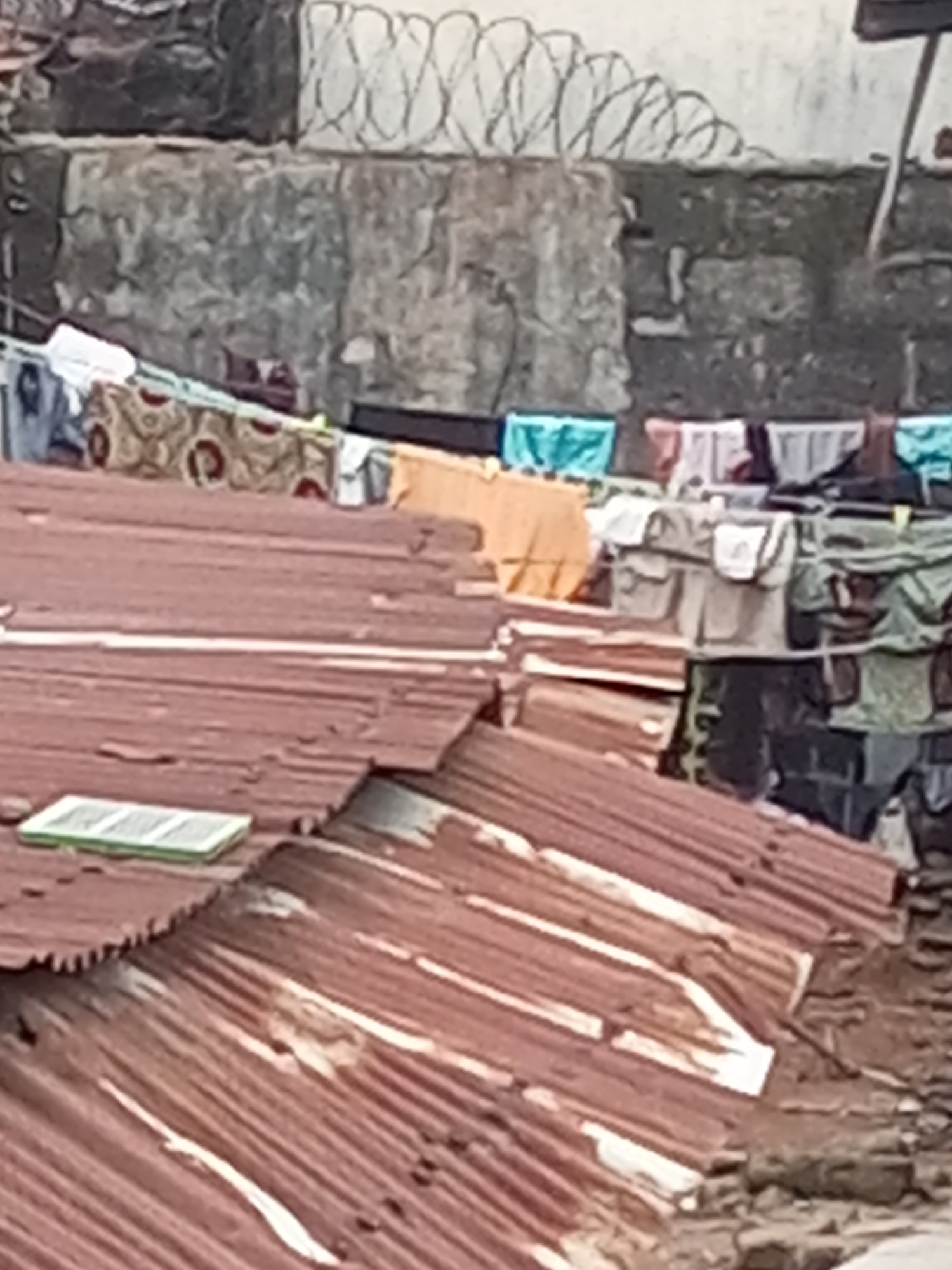 The image shows a corrugated iron roof in Onitsha, Nigeria. Laundry hangs on a line in the background, in front of a concrete wall topped with razor wire. The roof is rusted and dilapidated. There are no people visible.