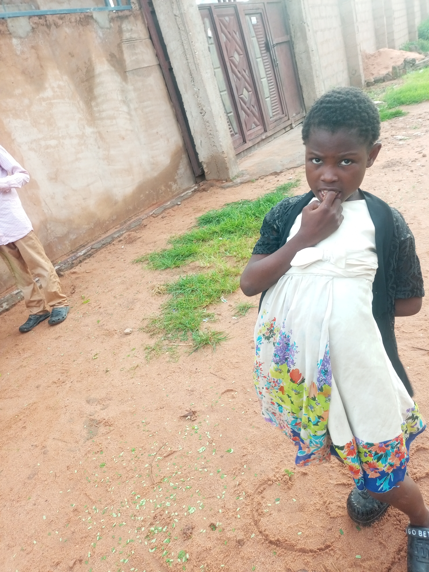 A young Black girl, with short, dark, curly hair, stands outside in a sandy area. She is wearing a light-colored dress with a floral pattern, a dark, lacy cardigan, and dark shoes. Her right hand is raised to her mouth, with her fingers near her lips. Her expression is serious. To the left of the girl, a person wearing a light pink and white striped shirt and tan pants is partially visible, with their feet in dark shoes. They are standing next to a textured, light brown wall. Behind the girl, a large, double metal gate with geometric patterns is visible, set within a concrete structure. The ground is sandy and scattered with small green leaves. The environment appears to be outdoors, possibly in an urban or semi-urban setting in Tempure, Nigeria, given the architecture and sandy ground.