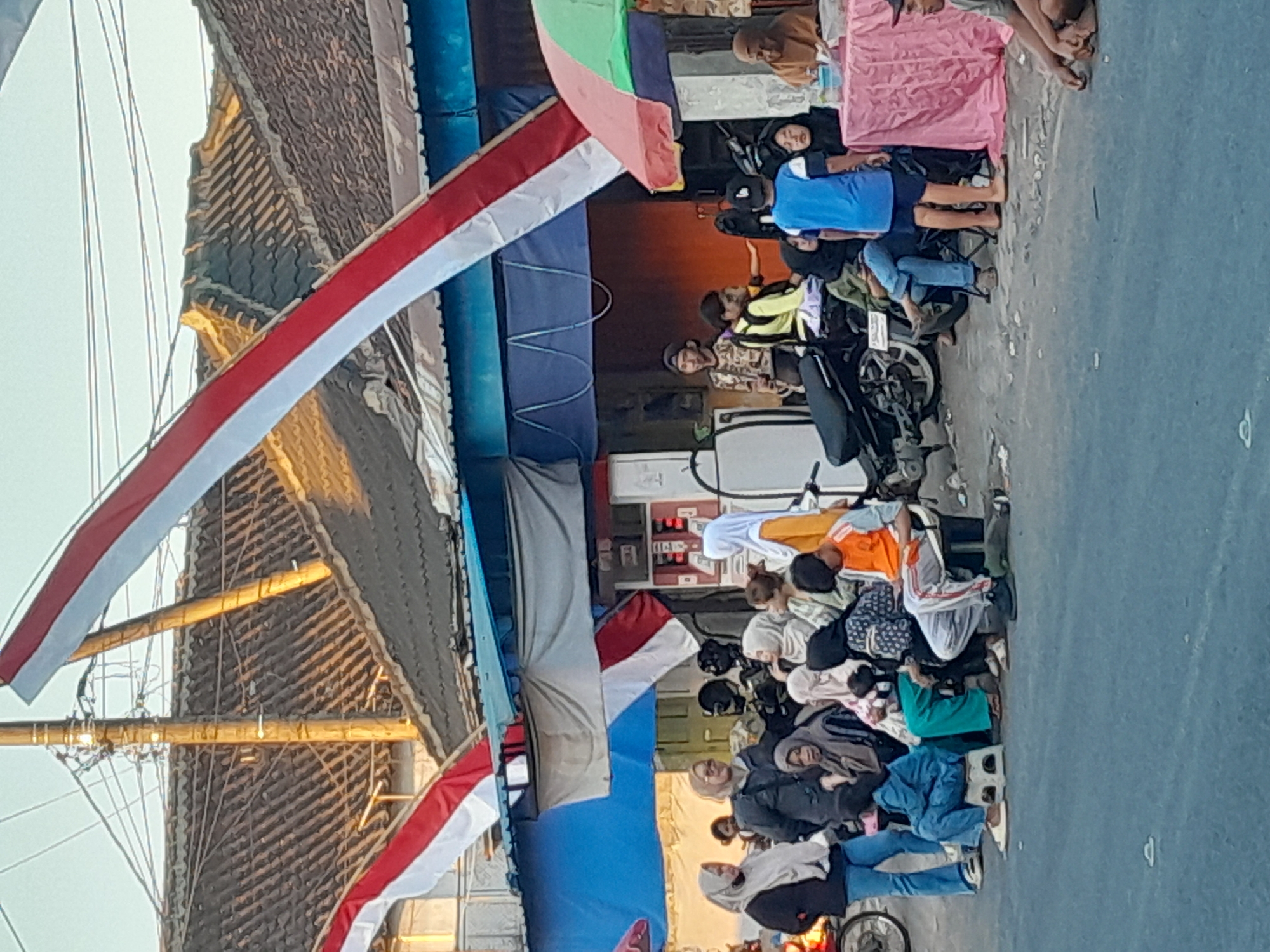 The image captures a street scene in Gentengwetan, Indonesia, with a group of people gathered near a building. Red and white Indonesian flags are draped across the foreground, suggesting a festive or nationalistic occasion. Several individuals, predominantly women wearing headscarves, are seated and standing, some appearing to be in conversation. Motorcycles are parked nearby, indicating a mode of transportation common in the area. A stall covered with a pink cloth is set up, with items on display. The environment appears to be a casual urban setting with a building featuring a tiled roof and electrical wires overhead. The lighting suggests it is daytime, possibly late afternoon due to the warm tones in the image.
