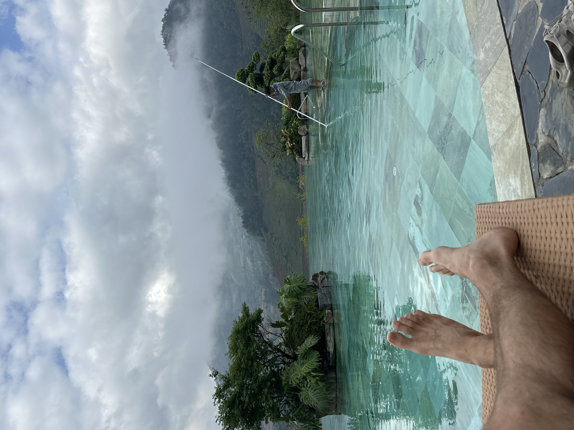 The image depicts a leisure scene at an outdoor infinity-edge swimming pool in Púng Luông, Vietnam, under a cloudy sky. In the immediate foreground, a person's bare feet are visible, resting on a light brown wicker lounge chair, indicating a relaxed posture. To the lower right, a pair of light gray sandals rests on dark, irregular stone paving next to the pool edge.
The pool itself features turquoise water with a checkered pattern of teal and gray-green tiles on its bottom, showing surface ripples. Two small, verdant islands are situated within the pool; the left island hosts a multi-trunked tree surrounded by large-leafed plants, while the right island features a smaller, manicured evergreen tree amidst rocks.
Mid-scene, a person, presumably a pool attendant, stands in the shallow end of the pool near the right side. They are dressed in light gray work attire, including a long-sleeved shirt and shorts, and are holding a long, white telescopic pole, actively engaged in cleaning or skimming the pool surface. Stainless steel handrails for pool entry are visible beside this individual.
The background reveals a expansive mountainous landscape characteristic of Púng Luông. The mountains are densely covered with green vegetation, including forests and visible terraced fields on the slopes. Thick, low-hanging clouds and mist partially obscure the upper reaches of the mountains, blending into an overcast sky with patches of brighter blue visible between the clouds.