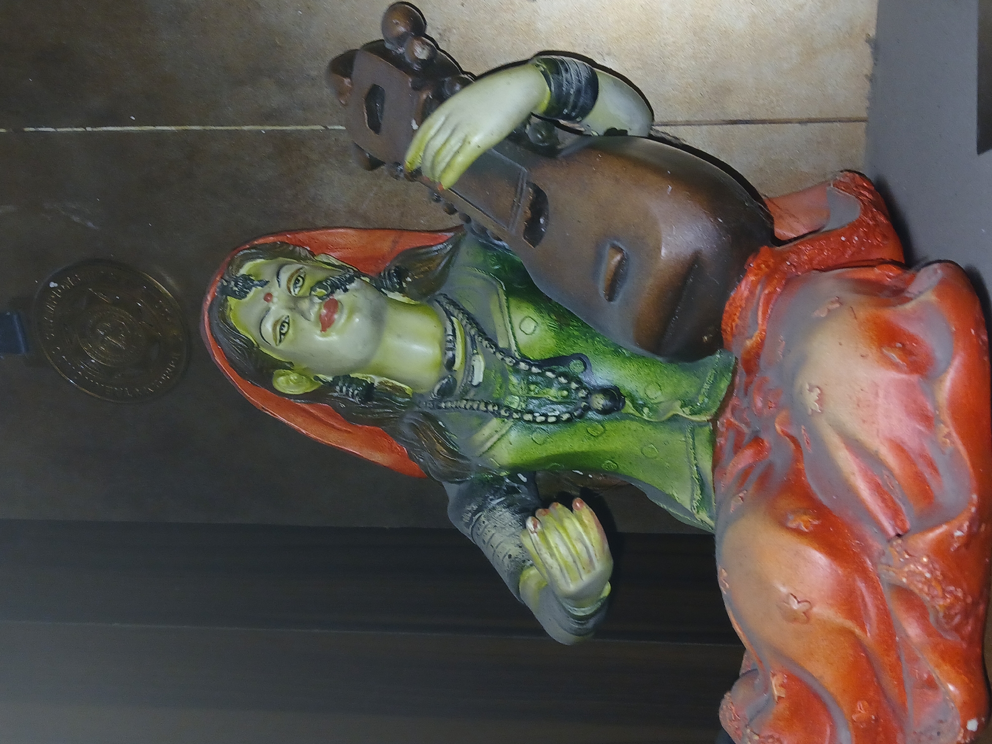 The image displays a vibrant, multi-colored statue of a woman playing a stringed musical instrument, possibly a sitar or veena. The statue, depicting a woman with light green skin, black hair, red lips, and a prominent red bindi on her forehead, is dressed in a green top and a draped orange and red lower garment. A black beaded necklace adorns her neck, and intricate details are visible on her wrists and feet. Her light-green hands are positioned on the brown instrument, suggesting she is actively playing it. Her feet, also light green with painted toenails, are visible at the bottom right.
The woman statue appears to be in a reclined or seated pose, looking upwards with a serene expression. The statue rests on a dark, textured surface, which could be wood or stone. In the lower-left portion of the image, a partially visible, round metallic object, resembling a large coin or medallion, lies on the same surface; any inscriptions or details on it are indistinct. The background is largely dark and out of focus. The photograph is taken from a slightly elevated angle, looking down at the statue, which is characteristic of traditional Indian artistry and decor, often found in places like Bengaluru, India.