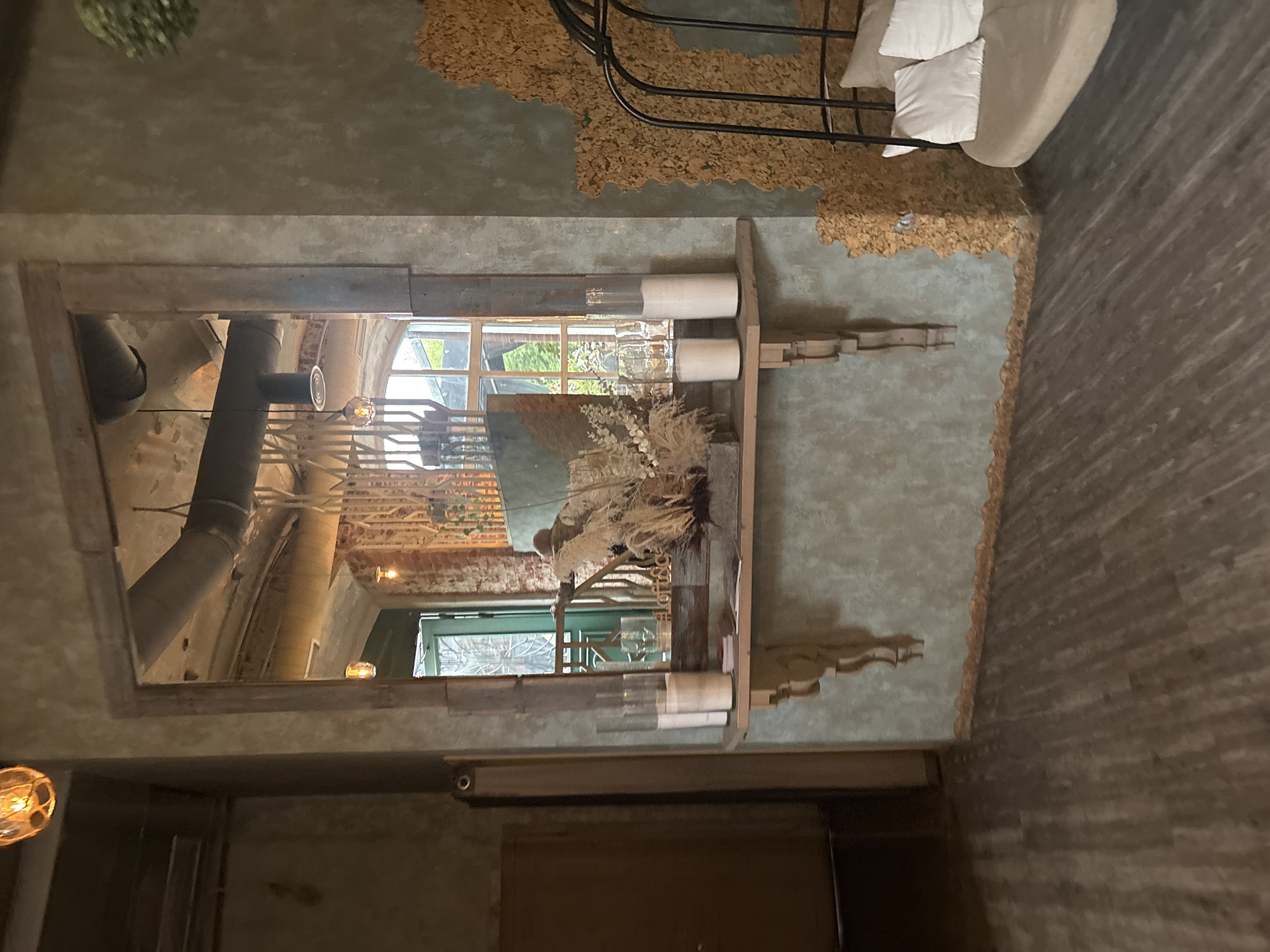 The image depicts an interior scene, likely a cafe or restaurant, with a rustic and industrial aesthetic. A large, wooden-framed mirror dominates the central portion of the image, reflecting a window with a green door and brickwork. On a shelf beneath the mirror are several glass cylinders containing white candles, and a decorative arrangement of dried grasses and reeds.
To the right of the mirror, a textured wall with a golden, cork-like material forms an interesting backdrop. Adjacent to this wall, a black metal hammock-like chair with beige cushions is visible. The floor is made of dark wooden planks.
Several light fixtures are visible, including an amber glass pendant light at the top left and another industrial-style pendant light with a circular shade reflected in the mirror.
The overall atmosphere suggests a cozy yet stylish establishment. The image does not contain any identifiable people. There is no specific indication of the location being in Ivanovo, Russia.