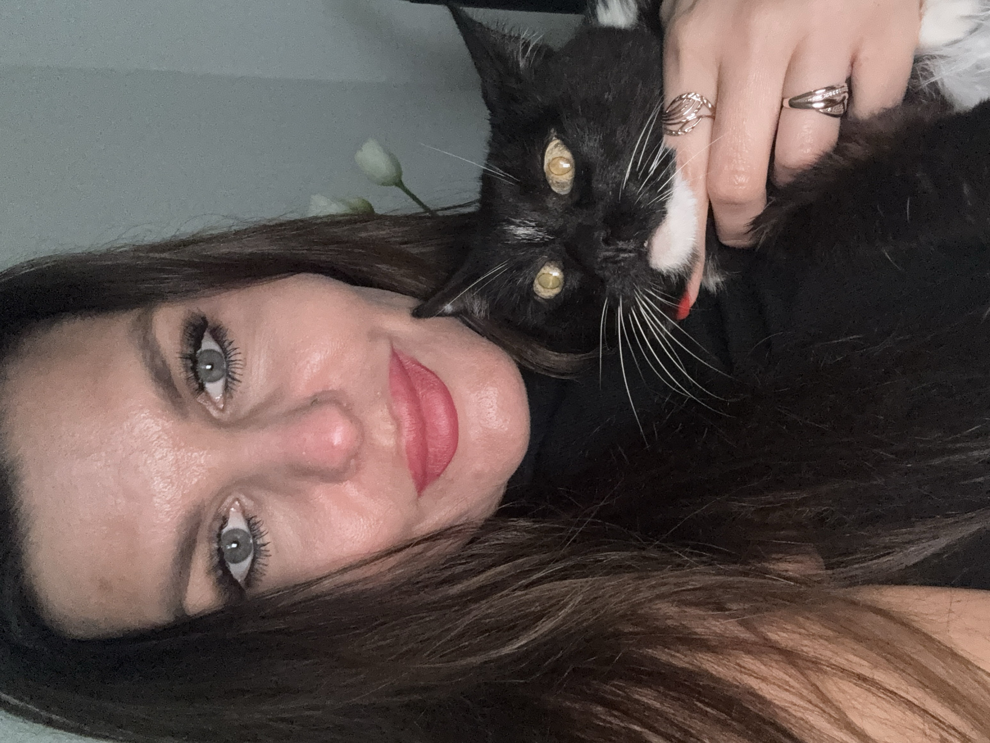 A close-up selfie captures a woman holding a black and white cat. The woman has long brown hair, grey eyes, and is wearing pink lipstick. She is wearing a black turtleneck. The cat has yellow eyes and is looking at the camera. The woman's hand is adorned with two rings. The background is a plain, grey wall with a hint of greenery in the upper right corner. There is no identifiable information about the location in the city of Chaikovsky, Russia.