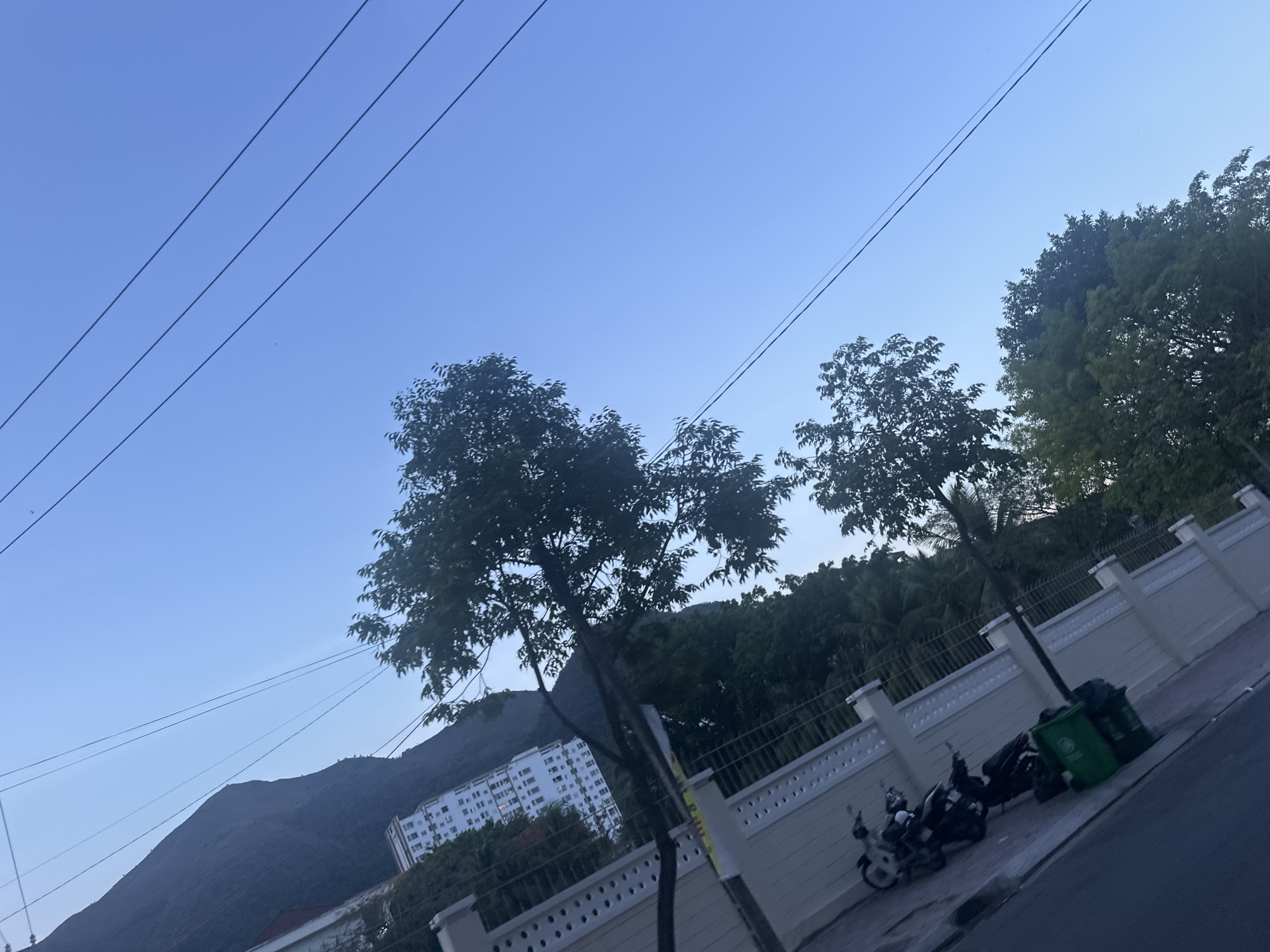 The image shows a street scene in Bac Nha Trang Ward, Vietnam. The sky is a clear blue, crossed by power lines. Trees line the street, and a white wall runs along the sidewalk. Several parked motorbikes and green trash cans are visible next to the wall. A building is visible in the background, nestled at the base of a mountain.