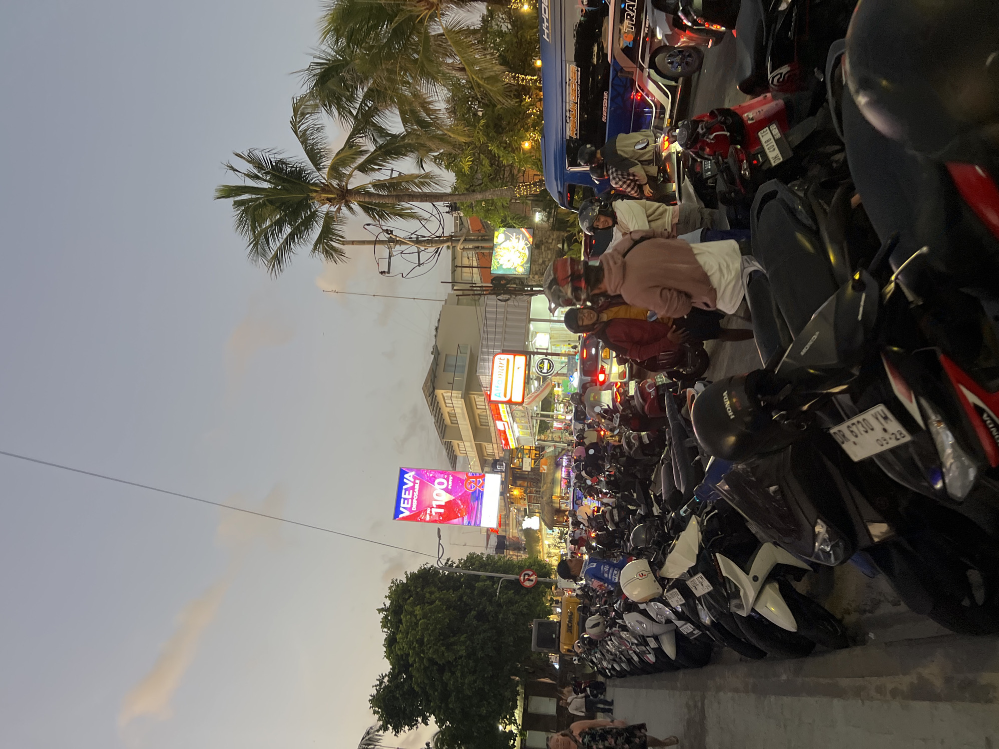 The image depicts a street scene in Kuta, Indonesia, at dusk. A long row of parked motorbikes lines the left side of the street. Several people, mostly young adults, are standing and milling about near the motorbikes, some wearing helmets. In the background, there are buildings with commercial signage, including a large digital billboard advertising "VEEVA". Palm trees are visible against the twilight sky, and a blue bus with lettering is in the middle ground. The atmosphere suggests a busy urban area, possibly a commercial or tourist district.