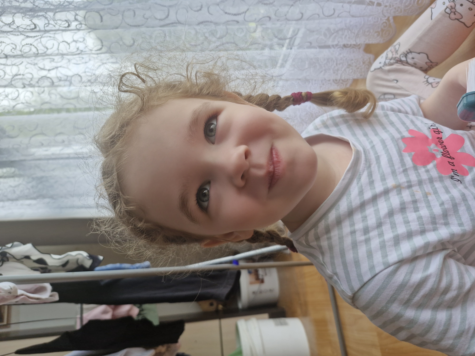 A young girl with blonde curly hair, styled in two braids, looks upwards and slightly to her right with a gentle smile. She is wearing a light gray and white striped t-shirt with a pink flower graphic and text that reads "I'm a flower girl". The background shows a white lace curtain behind her, and to her left, a metal drying rack with clothes hanging on it. In the further background, there are two white buckets and wooden flooring. The overall setting appears to be an indoor domestic environment, possibly a room in a house or apartment. There is no visible information to identify the specific location within Марковский, Russia.
