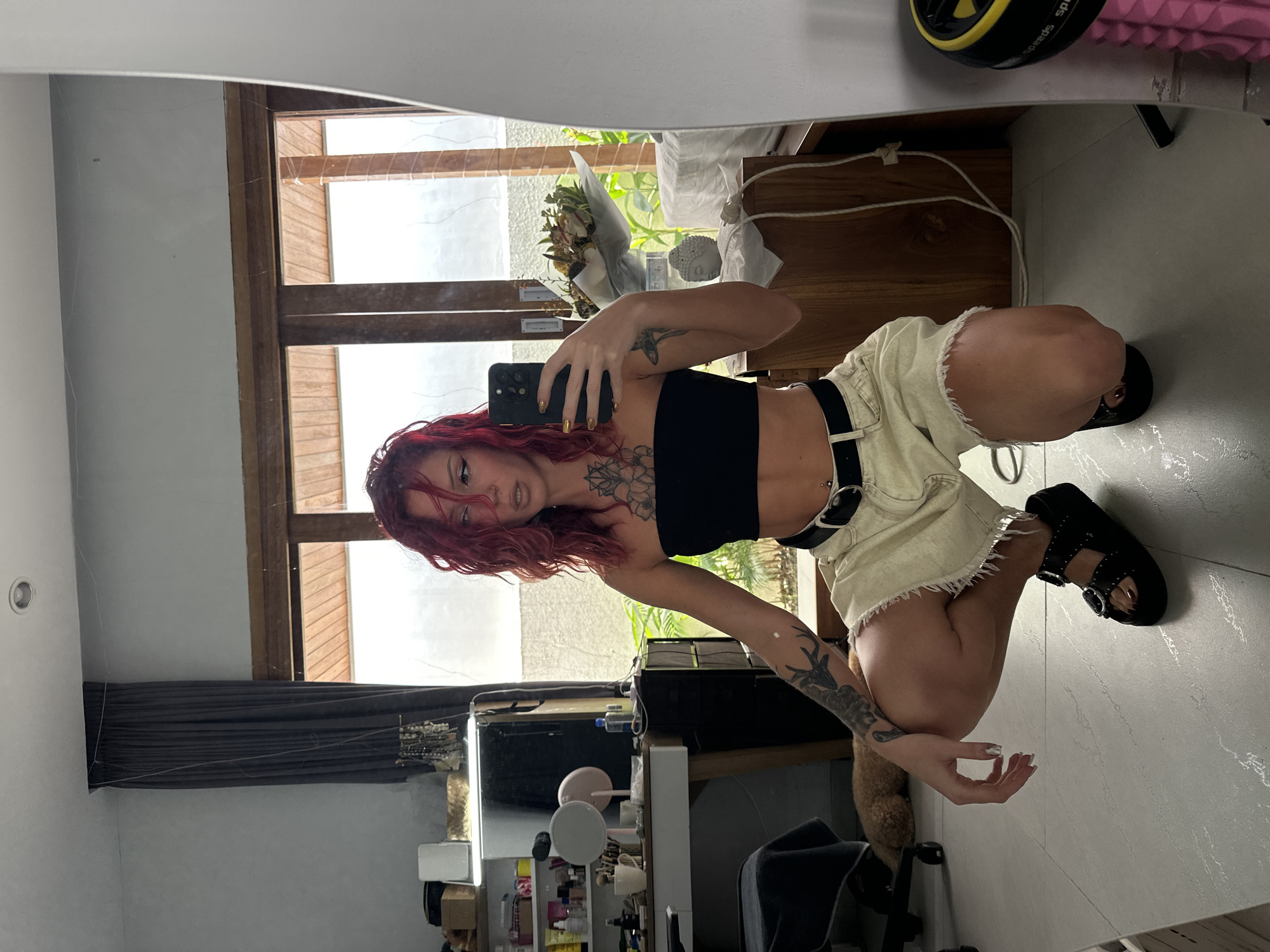 A red-haired female is photographed in a mirror selfie while kneeling on a tiled floor in a room. She is wearing a black tube top, light-colored shorts with a black belt, and black platform sandals. Tattoos are visible on her arms and chest. A black smartphone is held in her hand, capturing the selfie.
Behind her in the reflection, a window with wooden framing is visible, along with partially drawn grey curtains. A vanity with cosmetic products and mirrors is positioned to her left. Behind the window is an out-of-focus view of greenery. A bouquet of flowers is on a wooden dresser. A possible pet is standing near her legs.
The location is North Kuta, Indonesia.