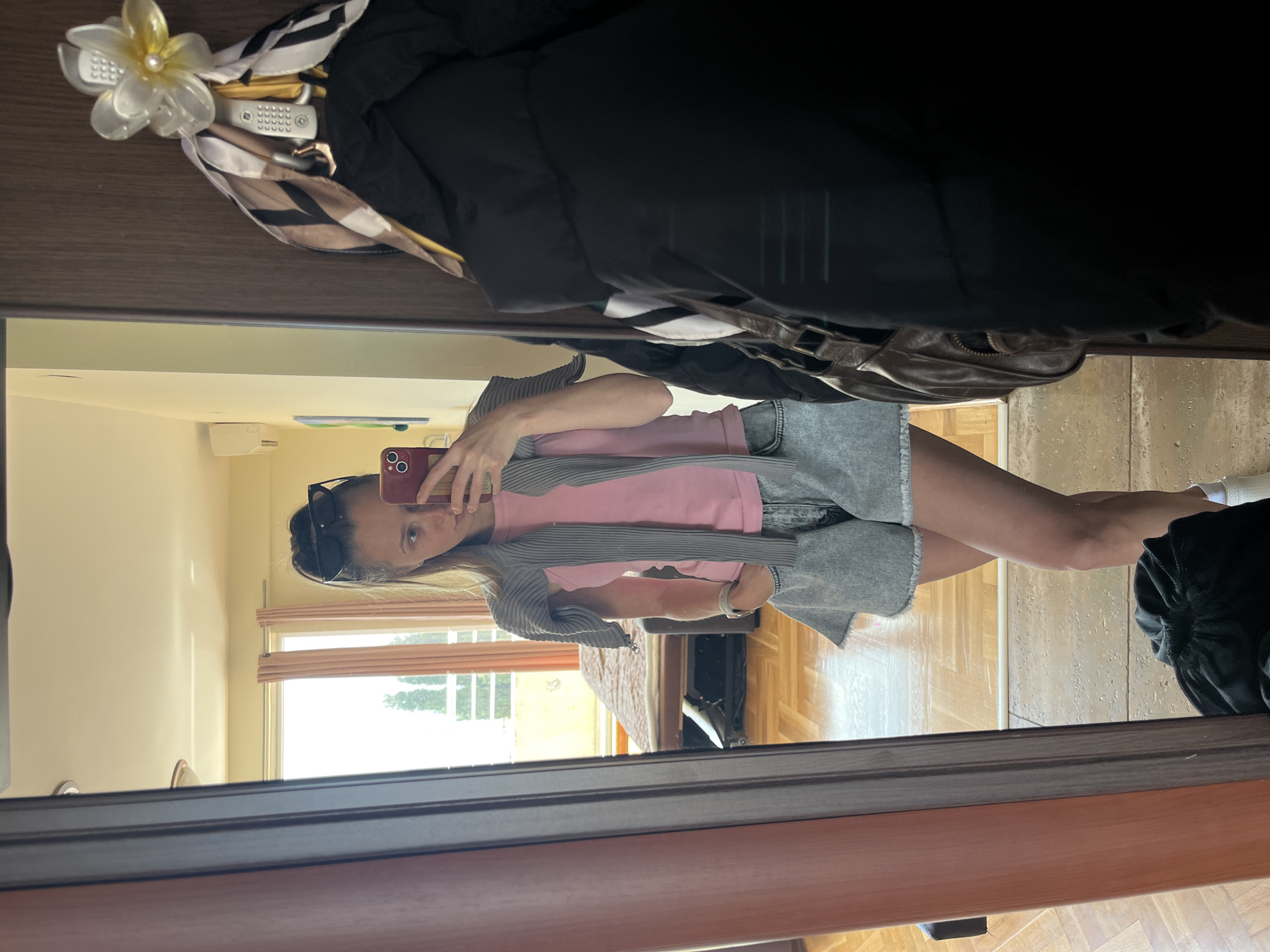 A woman is reflected in a mirror, holding a smartphone to take a picture of herself. She wears sunglasses on her head and has a pink t-shirt with a gray cardigan and a gray denim skirt. The room behind her has a window and light-colored walls. Overhead items, possibly coats or accessories, are visible at the top of the mirror. The room's flooring is a light wood parquet. The location is suggested to be in Bečići, Montenegro, based on provided context.