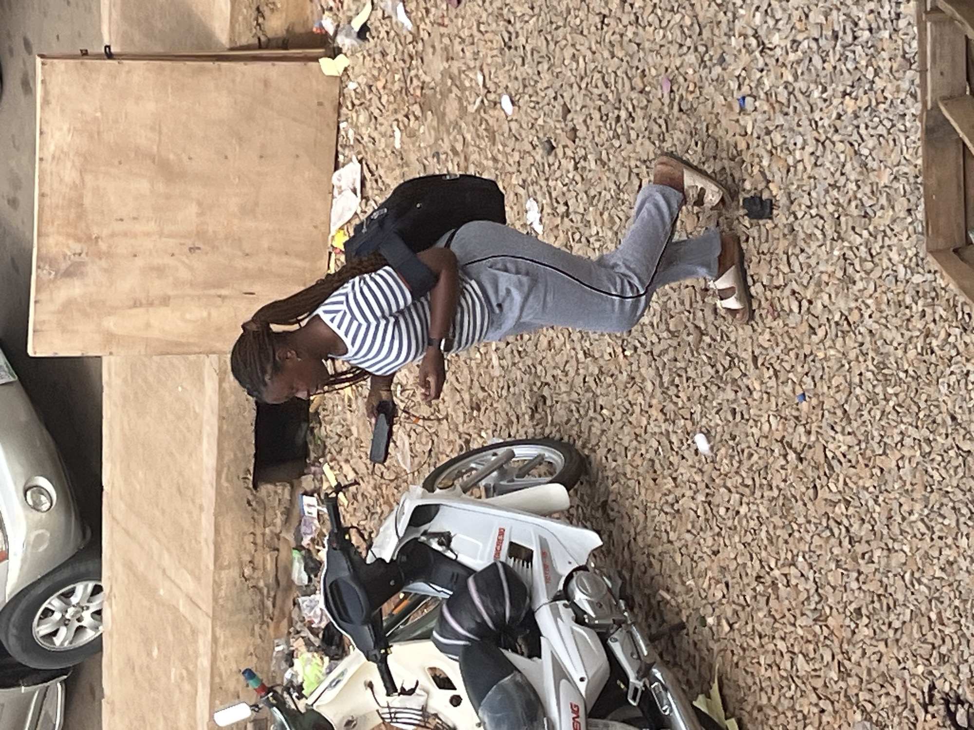 A young woman with braided hair is captured from a high angle, dressed in a striped t-shirt and grey sweatpants. She is holding a dark smartphone in her right hand. A black backpack is slung over her shoulders. She is standing on a ground covered with small, light brown pebbles. To her left, a silver car is partially visible, with its front wheel and fender showing. In the foreground, a white motorcycle is parked, with some of its parts covered by a black bag. The scene appears to be outdoors, possibly in an urban or semi-urban environment given the presence of vehicles and the material of the ground. The lighting suggests it could be daytime, with no specific weather conditions evident. The woman's expression and posture are neutral, and there are no obvious cues about the specific activity or event taking place, other than perhaps a brief pause. The setting is in Jos, Nigeria.
