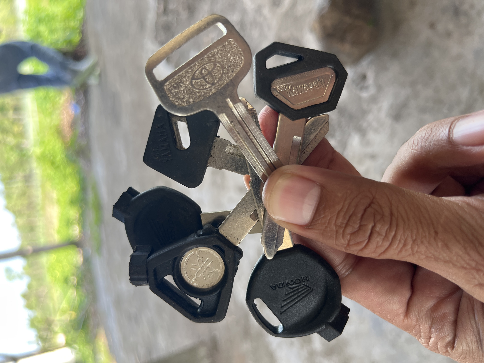 In the city of Kecamatan Pujut, Indonesia, a hand is holding a bunch of motorcycle keys. The keys have different brand markings including Honda, Yamaha, Kawasaki, and Toyota. The background shows a blurred outdoor scene with greenery and a person walking in the distance. The ground appears to be concrete or asphalt.