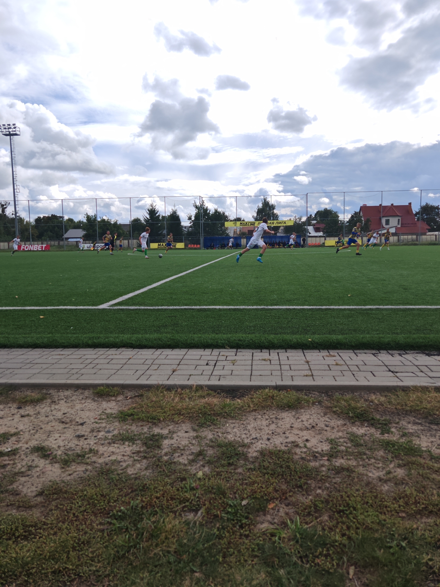 The image depicts a football match taking place on a green artificial turf field. Several young male athletes are visible on the field, actively engaged in playing the game. One player in a white uniform is dribbling a soccer ball, while others are running or positioned around the field. The background shows a fence, trees, buildings including a red-roofed house, and a cloudy sky with patches of blue. A prominent yellow banner with Cyrillic text, "MAXLINE ПРОВАЯ ЛИГА" (MAXLINE Premier League), is displayed behind the fence. On the left side of the field, another banner reads "FONBET". The scene appears to be an outdoor sports facility, likely a stadium or training ground within the city of Homyel, Belarus. The foreground consists of a paved walkway and patchy grass.