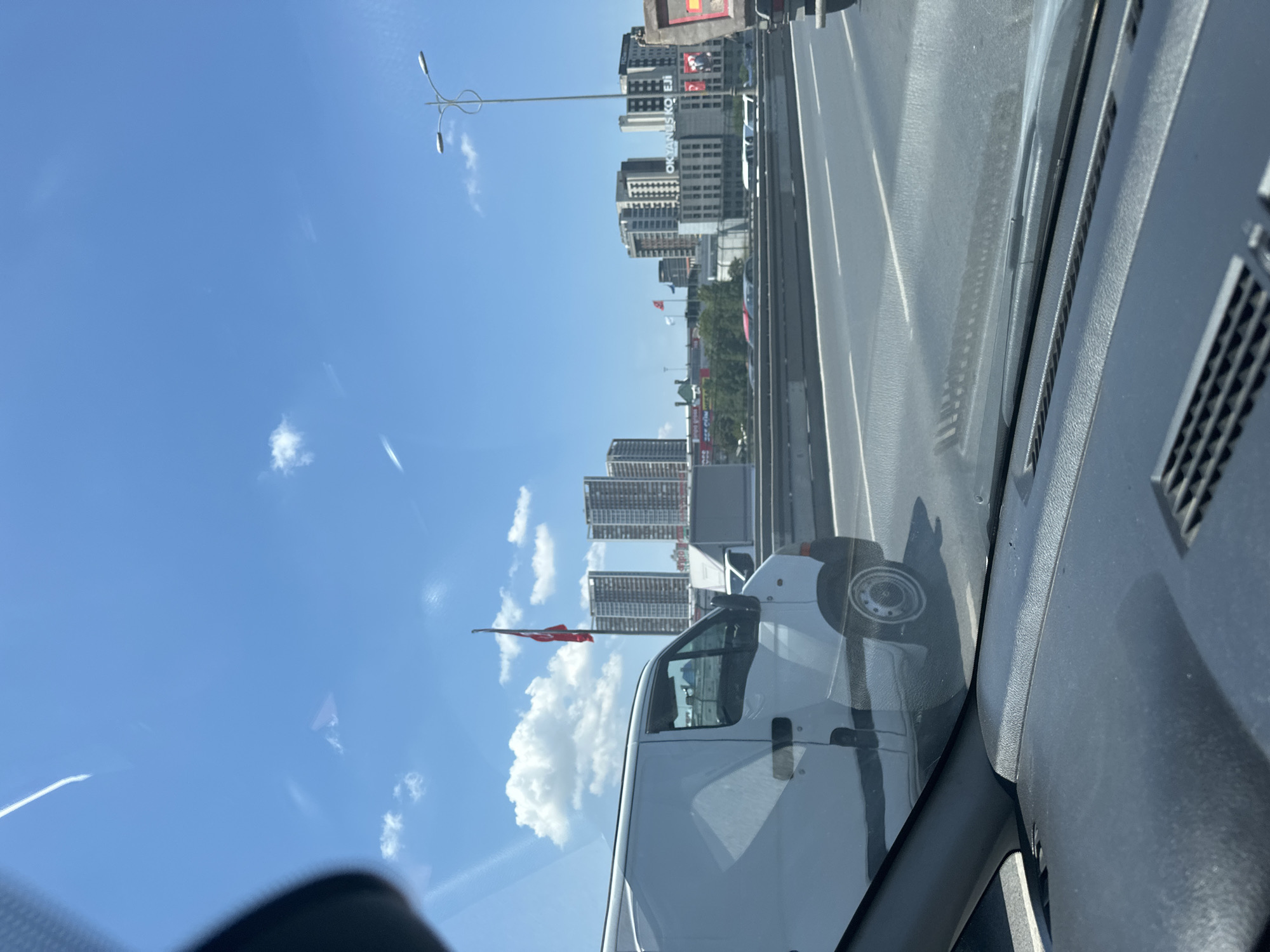 The image captures a daytime scene on a highway in Bayrampaşa, Turkey, likely during rush hour traffic. The viewpoint is from inside a vehicle, looking out through the windshield, which is partially obscured by the dashboard and a reflection of the sky.
Dominating the left side of the frame is a white panel van, its side facing the camera. The van appears to be in motion, suggested by the slight blur. The highway itself stretches across the middle ground, with multiple lanes indicated by white markings. In the distance, a cityscape rises, characterized by a cluster of modern, tall buildings. A prominent Turkish flag, red with a white crescent and star, is visible flying from a flagpole between two of the buildings.
Further in the background, other vehicles, including cars and what appears to be a truck, are visible on the highway and in the distance. A street light fixture with two lamps stands on the right side of the highway. The sky is a clear blue, with scattered white clouds and some thin streaks, possibly contrails. The overall impression is of a busy urban environment on a sunny day.