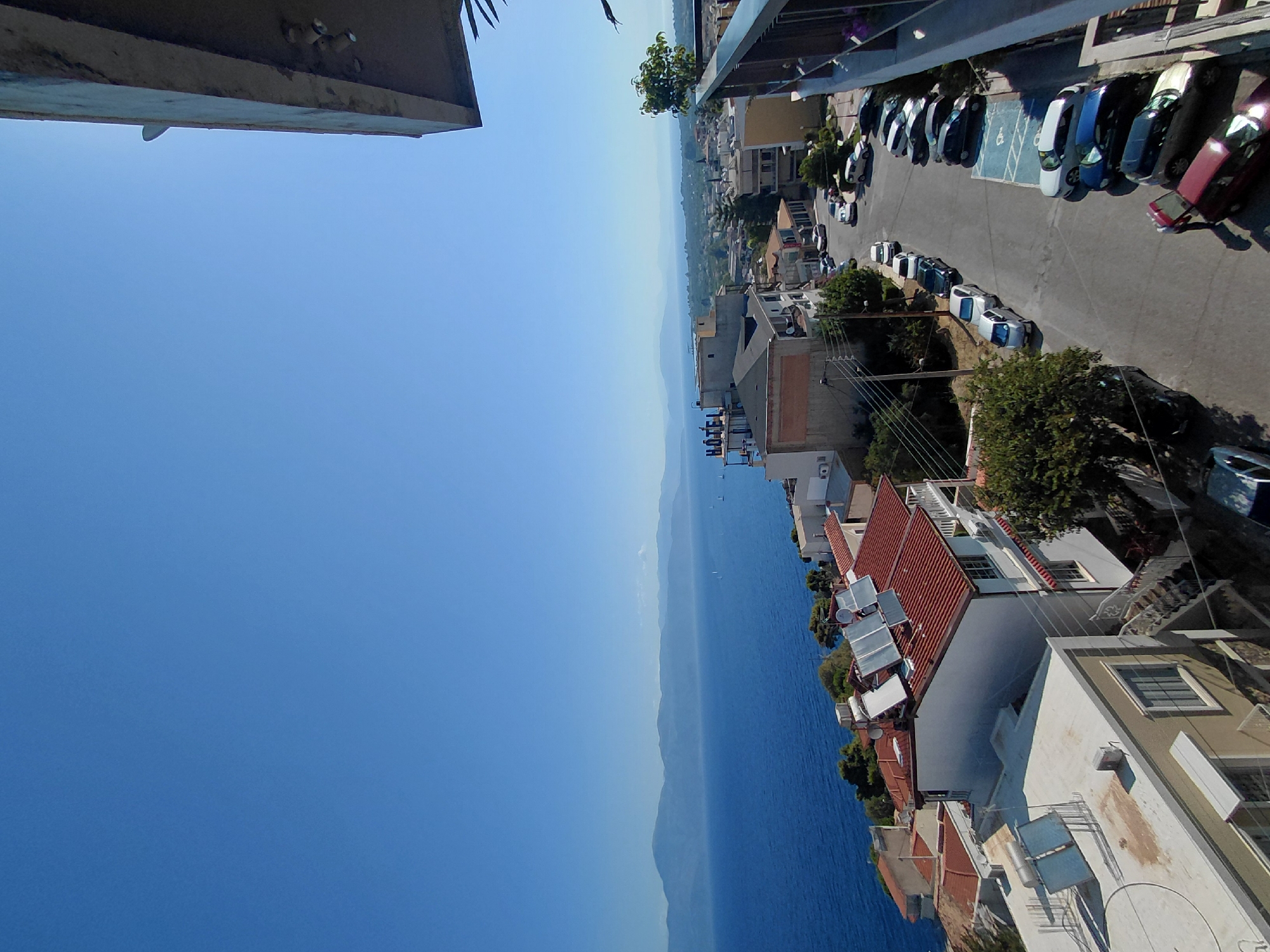 A scenic overlook shows a coastal town in the Municipal Unit of Aegio, Greece. The foreground features a balcony with a railing, overlooking a street lined with parked cars. Houses with red-tiled roofs and solar panels are visible, along with a building with the word "HOTEL" displayed prominently. Beyond the town, the deep blue sea stretches towards a distant mountain range under a clear, bright blue sky. The image captures a sunny day with a relaxed atmosphere.
