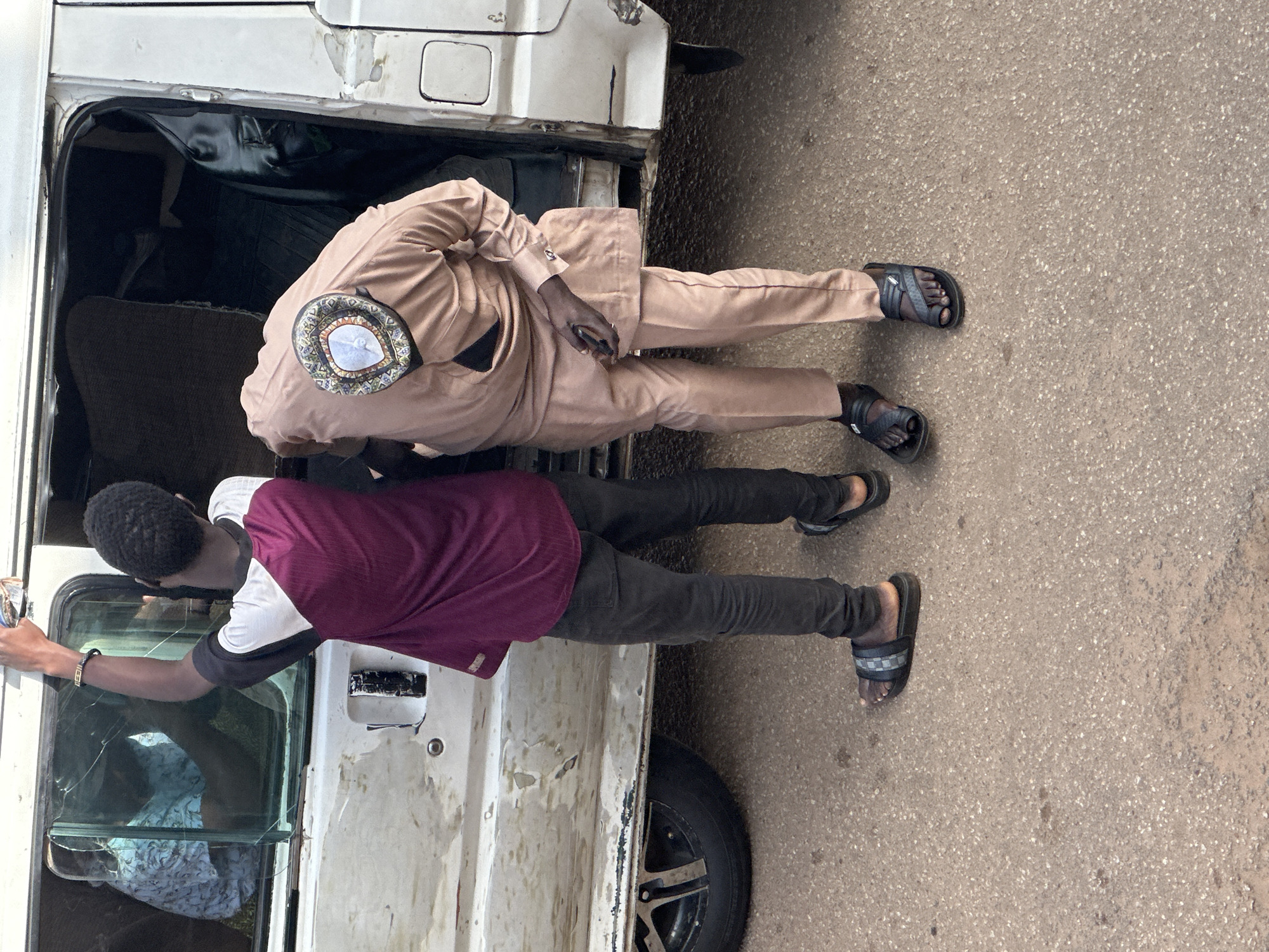 In the city of Bukuru, Nigeria, two individuals are positioned near the open door of a weathered, white van. The van's exterior displays signs of age and wear, with chipped paint and what appears to be a cracked window on the driver's side.
To the left, a young Black male, seen from the waist down, stands facing the van. He is wearing a maroon and white short-sleeved shirt, dark denim jeans, and black sandals with a checkered pattern. His right arm is raised, holding a small object, possibly a cleaning rag, as he appears to be cleaning the van's window.
To the right and slightly in front of him, another Black male is bent over, reaching into the van. He is dressed in a tan colored traditional outfit, consisting of a tunic and matching trousers, accessorized with a decorative cap adorned with a circular, embroidered pattern. He is wearing dark sandals. His posture suggests he is retrieving or placing something inside the van.
The ground is a rough, asphalt-like surface, indicative of a street or roadside. The lighting suggests it is daytime.