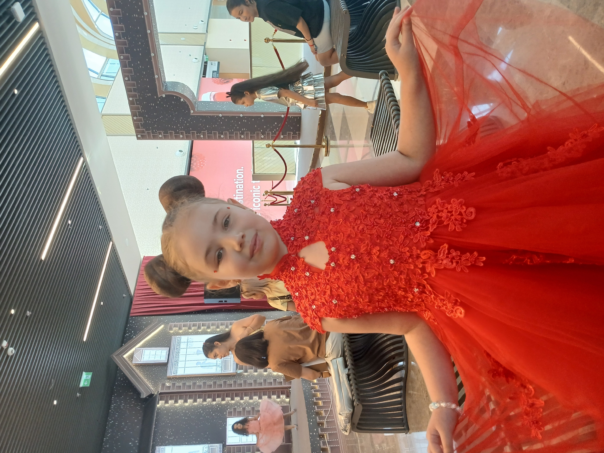 A young girl in a red, sequined dress with her hair in two buns is looking at the camera. Her dress has a floral pattern on the bodice. She is sitting in front of a backdrop with a city skyline design. To her left, a woman in a silver dress is posing. Behind her, another woman in a black shirt and white shorts is watching. In the background, there is a large red banner with text that is partially visible. The setting appears to be an indoor event or photoshoot.