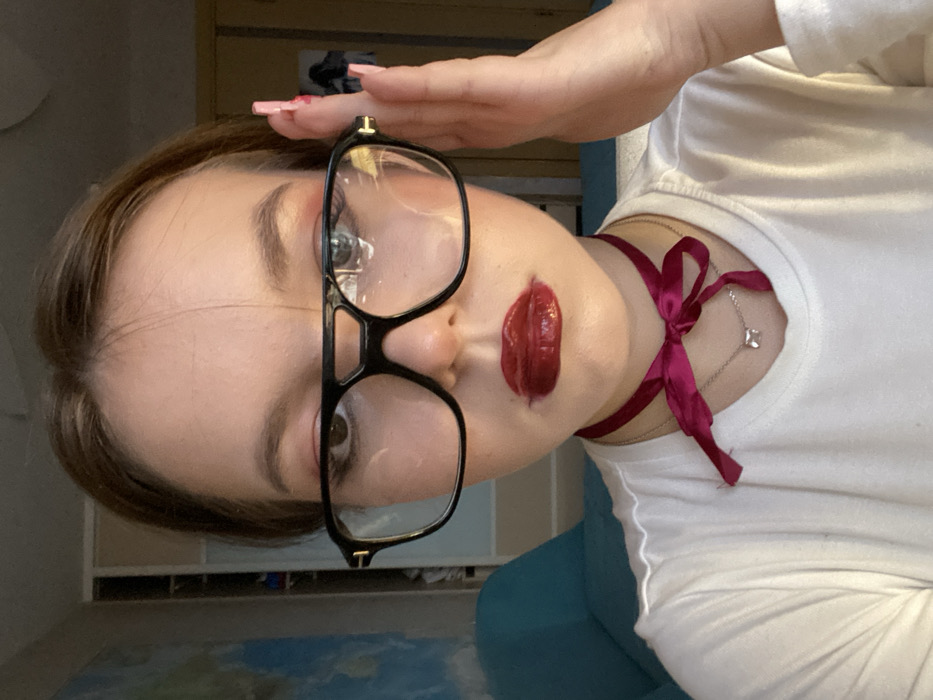 A close-up selfie captures a young individual, appearing female, with light brown hair, fair skin, and brown eyes. The subject wears large, black-framed glasses with clear lenses, featuring a double bridge and gold-colored accents on the temples. Makeup includes visible pinkish-red eyeshadow and dark red, highly glossy lipstick. A dark magenta satin ribbon is tied in a bow around the neck, situated above a delicate silver-colored chain necklace with a small, clear, rectangular pendant. The subject's right hand, displaying long, pointed fingernails painted light pink, is raised to the right temple of the glasses. The background is a blurred indoor environment. On the left, a large light blue area, possibly a map or mural, is partially visible, alongside a teal piece of furniture. On the right, a light-colored wall features a vertical seam or door frame, and indistinct dark items are visible in the far background. The image does not contain identifiable markers indicating a specific location within the city of Chaikovsky, Russia.