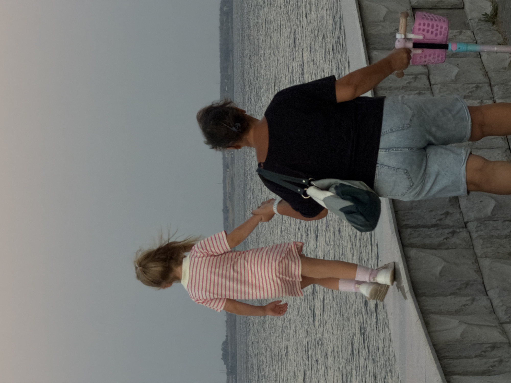 An adult woman and a young girl are walking along a concrete embankment next to a large body of water, possibly the Volga River, in Kamyshin, Russia. The girl, facing away from the viewer, wears a pink and white striped dress and pink socks. She holds the woman's hand. The woman, also facing away, wears a black t-shirt and denim shorts. She carries a bag over her shoulder and holds the handlebars of a pink and blue scooter. The sky is overcast.