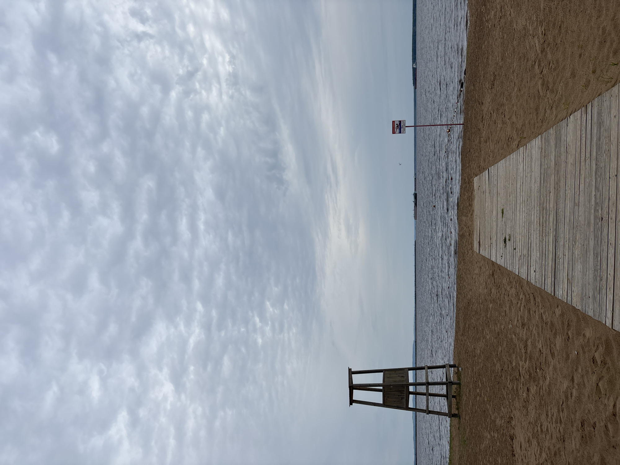 This vertically oriented image captures a serene, overcast day at a beach or lakeside in Chaikovsky, Russia. The majority of the left side of the frame is dominated by a pale gray sky filled with textured, streaky clouds, suggesting an overcast or partly cloudy weather condition.
On the right side of the image, divided horizontally, is a body of water (likely the Kama Reservoir) in the upper portion and a sandy shoreline with a wooden path in the lower portion. The water's surface is lightly rippled, indicating a calm state, and a distant, faint landmass or shoreline is visible on the horizon. A line of small, dark buoys or markers stretches across a section of the water, likely delineating a swimming or restricted area.
Along the sandy beach on the right, a weathered wooden plank path extends diagonally into the frame from the bottom-right corner. Closer to the water's edge, towards the center-right of the image, a red pole stands with a rectangular warning sign attached. The sign features a red border, a white background, and clearly displays a red circle with a diagonal line over a figure in water, universally recognized as a "No Swimming" symbol. While the accompanying text is small, it likely reinforces "Купаться запрещено!" (Swimming forbidden!).
Towards the bottom-left of the image, situated on the sand near the water, is a rustic wooden structure resembling an elevated lifeguard chair or observation stand. No people are visible anywhere in the scene.
The lighting is soft and diffused due to the heavy cloud cover, making the precise time of day ambiguous, though it appears to be daytime. The overall atmosphere is quiet and somewhat tranquil, devoid of typical beach activity.