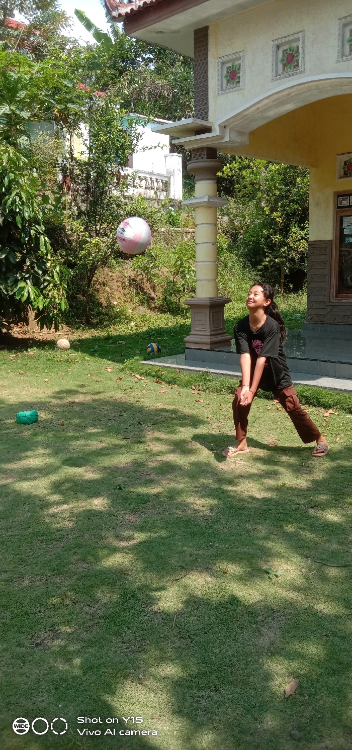 The image depicts an outdoor scene in Ranugedang, Indonesia, featuring a young female individual interacting with a ball. She is positioned slightly right of the frame's center, dressed in a dark t-shirt, brown cargo-style pants, and light-colored flip-flops. Her body is in a low, bent-knee stance with arms joined, typical of a volleyball pass, and her head is tilted back, looking upwards with a smiling expression. A pink and white striped ball, bearing the text "OFFICIAL," is suspended in mid-air directly above and slightly in front of her.
On the grassy ground, a light-colored, possibly deflated, ball rests to the left of the central action, and a yellow and blue striped ball is situated further back near the base of a house structure. A small green, bowl-shaped object is visible on the grass in the foreground, left of center.
The right side of the image features a light-colored house or building structure. It displays an arched entryway or overhang supported by a prominent pillar with horizontal brown textured accents and decorative square wall panels containing floral designs. A dark-framed window is visible on the far right, and the lower wall features dark gray tiling. The background consists of dense green foliage, including trees and shrubs, with glimpses of distant white building structures and balustrades. The foreground is covered in lush green grass marked by distinct patches of sunlight and shadow. The sky is clear, indicating bright daylight.