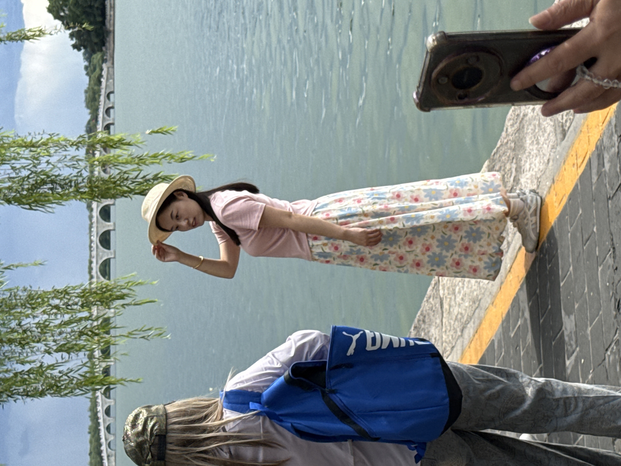 This image depicts an outdoor scene at the Summer Palace (Yiheyuan) in Haidian District, Beijing, China, identifiable by the distinctive Seventeen-Arch Bridge visible in the background across Kunming Lake. In the foreground on the left, a person with long, light-colored hair, wearing a camouflage baseball cap, a light purple long-sleeved top, and patterned grey trousers, stands with their back to the camera. They are carrying a bright blue backpack with a partially visible white "Puma" logo and text.
Centered in the middle ground, a young East Asian woman with long dark hair, wearing a light pink t-shirt, a long white skirt with a blue and pink floral pattern, and light-colored sneakers, stands near the water's edge. She is holding a straw hat slightly above her head with her right hand and has a gold-colored bracelet on her left wrist. She is looking slightly to her left, posing for a photograph.
On the far right, a hand is visible holding a dark-colored smartphone with a prominent circular camera module, vertically oriented as if taking a picture of the posing woman. A beaded ring or phone accessory is attached to the phone case.
The background features the calm, greenish-blue water of Kunming Lake. Across the lake, the white stone Seventeen-Arch Bridge stretches horizontally, with green trees lining the distant shore behind it. Overhanging willow branches with long green leaves descend into the upper portion of the frame. The foreground shows a paved walkway of grey rectangular pavers, marked by a bright yellow line along its edge where it meets the water.