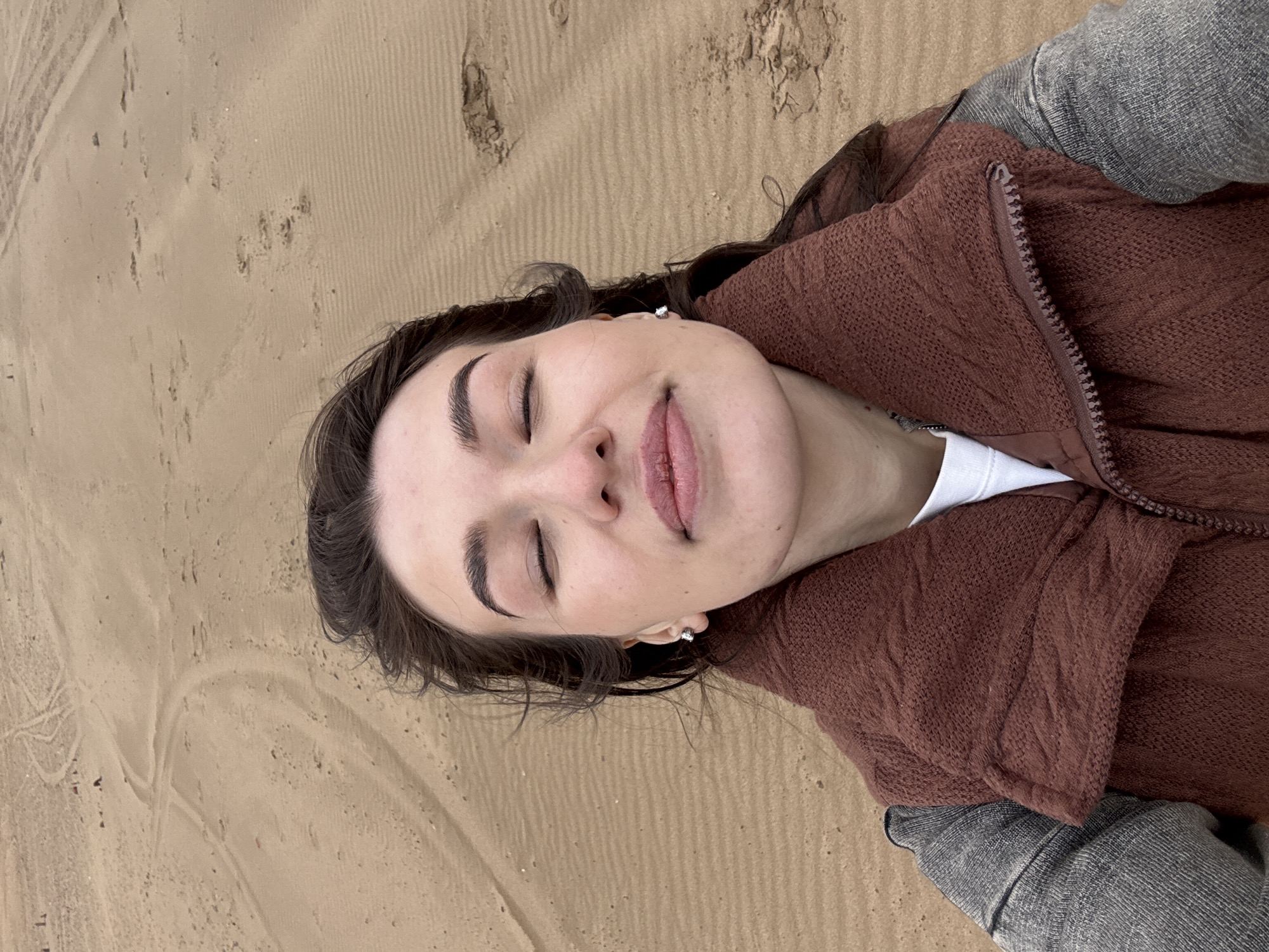 The media file captures a close-up, overhead view of a person lying down horizontally on sandy terrain. The subject is a young individual with dark hair, dark eyebrows, and a fair complexion. Their eyes are closed, and their lips are slightly parted with a hint of light pink lip color, conveying a serene and relaxed expression. A small, shiny earring is visible in their right ear.
The person is dressed in a textured, brown or maroon knitted jacket with a visible zipper. Underneath, a white top is peeking out. Portions of grey or blue fabric, likely pants or sleeves, are visible towards the bottom right of the frame, complementing the jacket.
The setting is an outdoor sandy environment, possibly a beach, dune, or a sandy area near a body of water. The sand exhibits various patterns and textures, including distinct parallel lines that could be from tire tracks, a rake, or natural wind ripples, along with scattered small footprints or irregularities. The lighting appears soft and natural, suggesting a daytime scene, possibly under an overcast sky or during the gentler hours of morning or late afternoon, contributing to the tranquil mood. No text is visible in the image. This scene is set in Noviy, Russia.