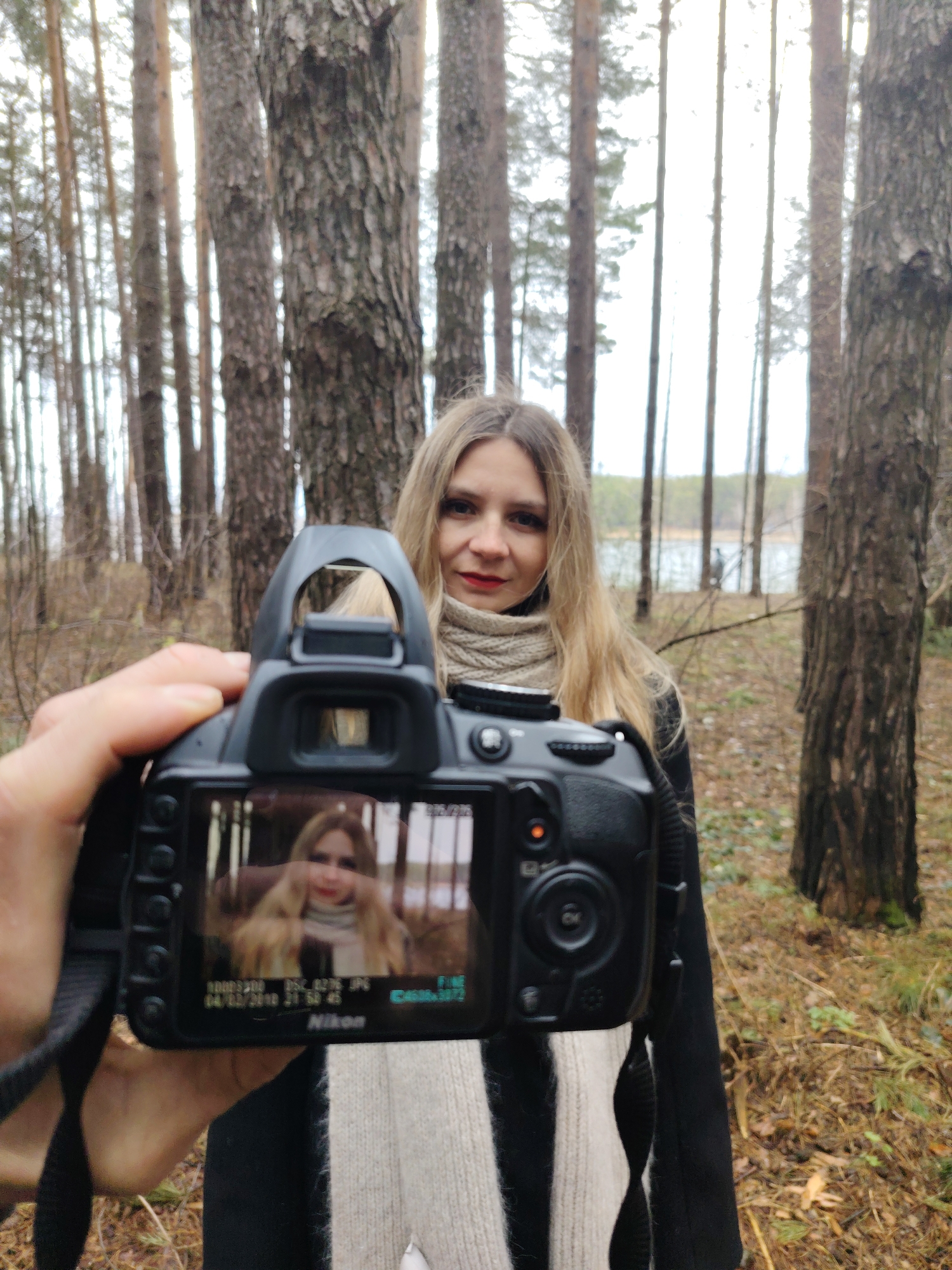 The image depicts an outdoor photography session set in a forest near Chaikovsky, Russia, during what appears to be autumn or early winter. The scene is captured from the first-person perspective of a photographer holding a camera.
In the left foreground, a hand holds a black Nikon DSLR camera, pointed towards a woman. The camera's rear LCD screen is prominently visible, displaying a live preview or a captured image of the subject, showing her reflection. Visible text on the camera screen includes "Nikon," "100D3200," "FXC 0036.JPG," "FINE," and a timestamp "04/12/2018 21:50:43," along with other camera settings.
The main subject of the photograph is a woman with long, blonde hair, who is positioned in the midground. She is wearing a light-colored, possibly beige, knitted scarf wrapped around her neck and a dark coat. She looks towards the viewer with a soft, slightly smiling expression, her face showing subtle makeup including red lipstick.
The background features a dense forest composed of numerous tall, slender trees with rough bark, characteristic of pine or fir trees. Through the trees, a body of water, likely a lake or river, is visible under a bright, overcast sky. The forest floor is covered with dry leaves and sparse, brown vegetation, indicating the season. The lighting is soft and diffused, typical of an overcast day, creating a gentle and natural ambiance. The image effectively portrays the act of photography by showing both the photographer's tool and the subject within the same frame.