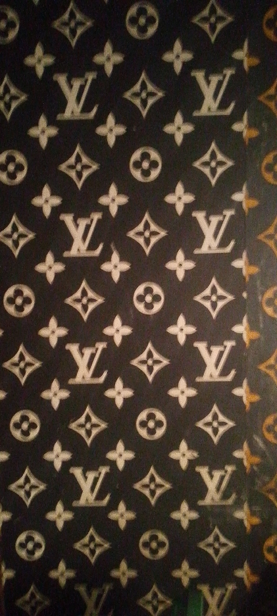The image shows a close-up of a fabric with a repeating pattern of Louis Vuitton motifs. The pattern consists of the Louis Vuitton "LV" monogram and the four-petal flower symbol, interspersed with diamond shapes containing four petals. The background is dark, likely black, and the motifs appear to be a lighter color, possibly white or a pale gold, with some metallic sheen. The lighting creates a slight sheen and some shadow effects on the fabric. The overall impression is of a luxury branded textile. There is no identifiable location in the city of Jalingo, Nigeria depicted in this image. The image is solely focused on the fabric pattern.