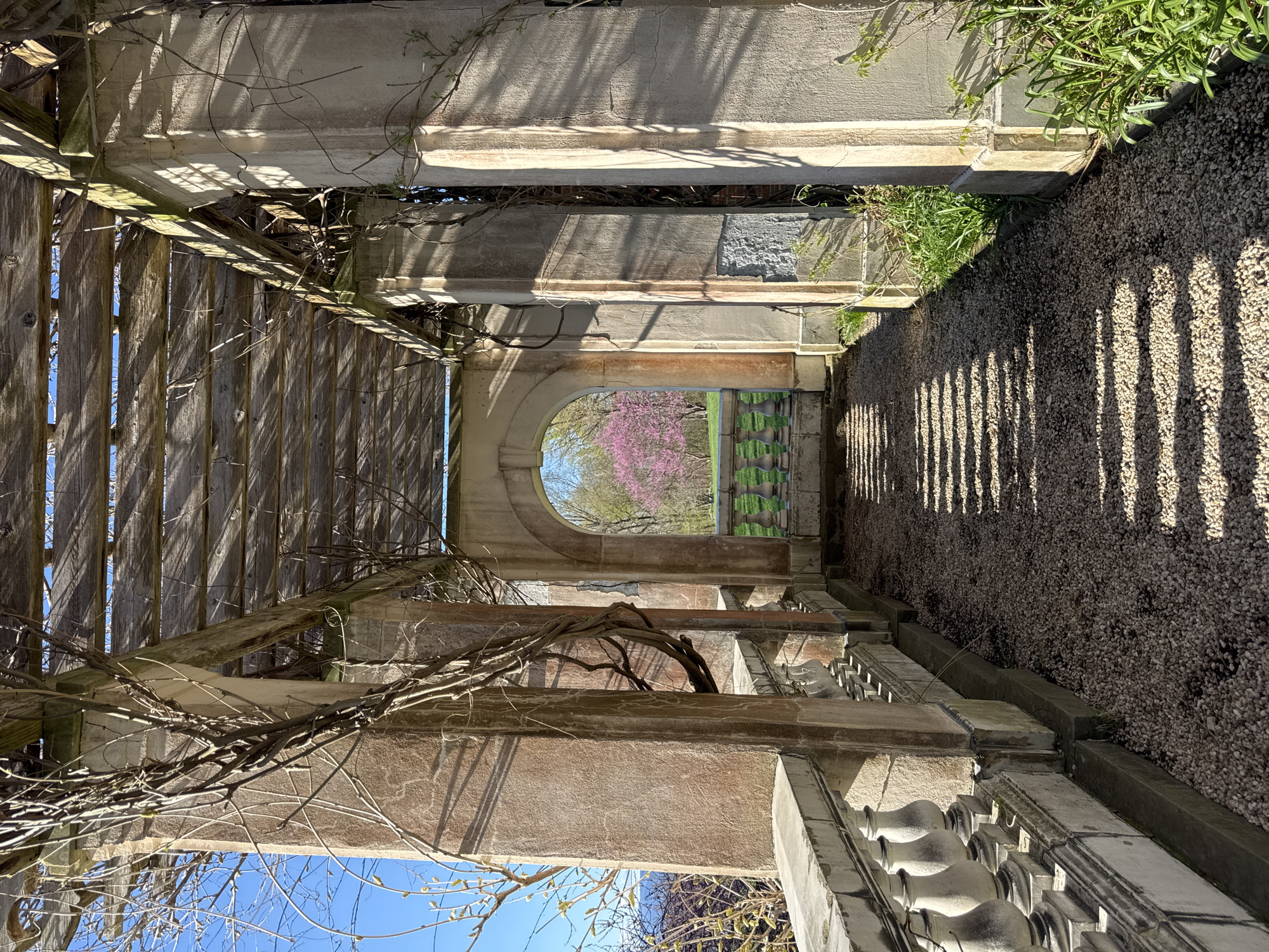 The image captures a view through an architectural structure located in the Town of Red Hook, United States of America. The structure consists of weathered stone and wood elements, including a pergola-like roof with wooden slats casting shadows on the stone pathway below. An arched opening in the structure frames a view of trees, including one with pink blossoms. The path appears to be covered with small pebbles or gravel. Bare vines are intertwined with the structure. The image is taken from a perspective that emphasizes the architectural elements and the landscape visible through the opening.