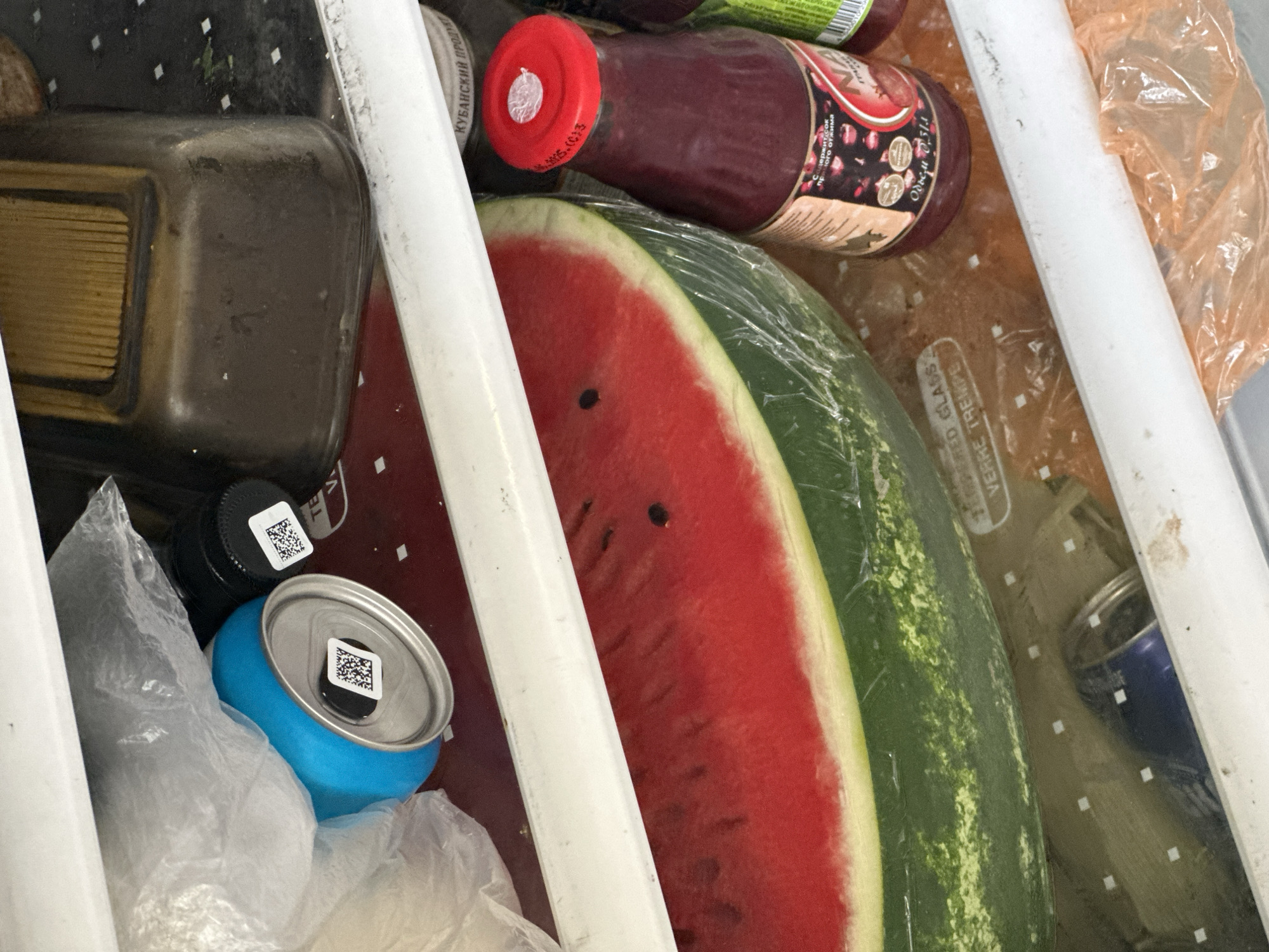 The image depicts the inside of a refrigerator shelf. A large slice of watermelon, wrapped in plastic, is prominently displayed. To its right, a bottle of dark red liquid, possibly juice, with a red cap and label featuring Cyrillic text, is visible. Above the watermelon, a blue can and a black-capped bottle with QR codes are partially obscured by a plastic bag. In the background, a brown, ribbed container sits on a shelf. The overall scene suggests a domestic setting, likely in Nevinnomyssk, Russia, given the Cyrillic labeling.