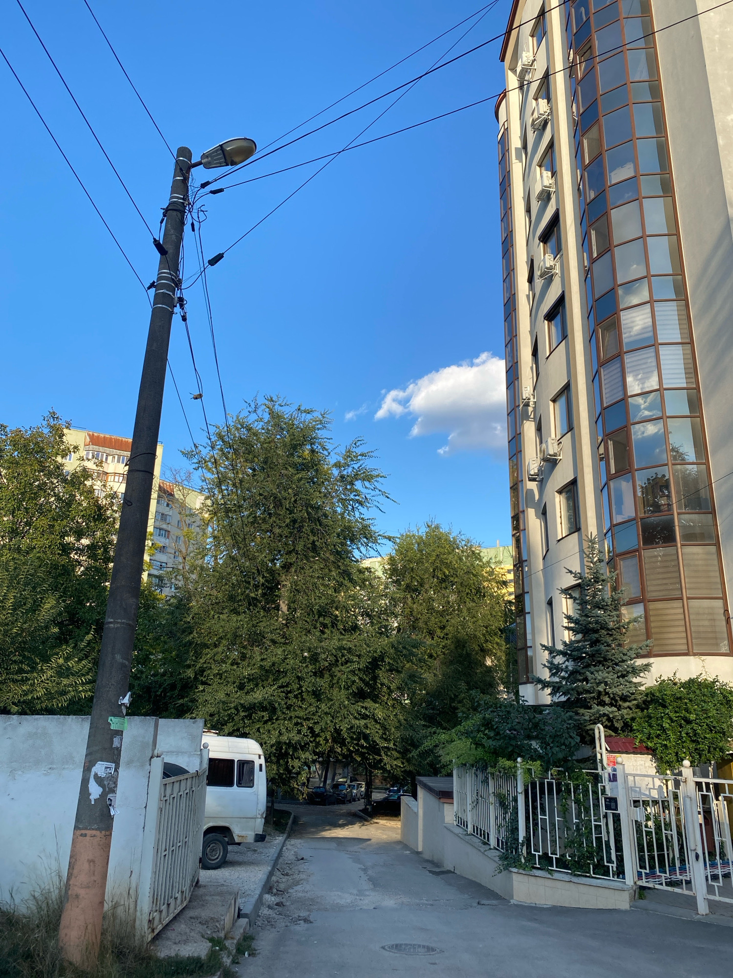 This is an exterior, eye-level, medium shot on a sunny day of a residential street in Chișinău, Moldova. On the left, a grey utility pole with a streetlamp is positioned near a white concrete wall covered in graffiti. Behind it, a white minibus is parked. Further back, several multi-story apartment buildings with beige and light green facades can be seen.
On the right, a modern, tall residential building with a curved facade of tinted glass windows dominates the scene. Several air conditioning units are visible on its exterior. Lush green trees and bushes, including a small fir tree, are planted in front of this building, separated from the street by a white metal fence.
The sky is a clear blue with a few scattered white clouds. The overall scene depicts a mix of older and newer residential architecture in an urban setting.