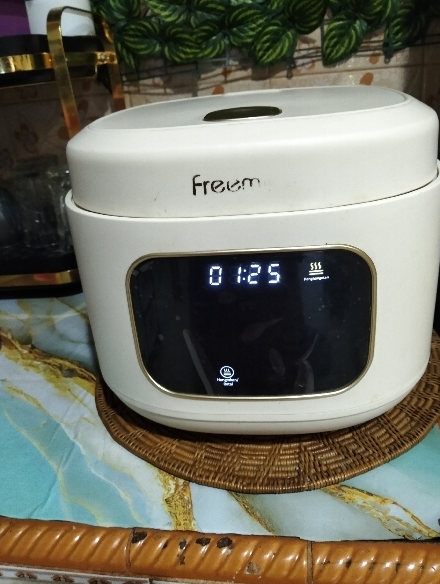 A close-up, eye-level shot shows a white rice cooker on a woven placemat. The rice cooker has a digital display showing "01:25" and an icon with three wavy lines indicating "Penghangatan" (Heating). To the left of the display, there is a button labeled "Hangatkan/Batal" (Warm/Cancel). The rice cooker has the brand name "Freem" printed on its lid. In the background, a blurred image of a shelf with greenery and kitchen utensils is visible. The scene appears to be in a domestic setting, possibly a kitchen in Muncar, Indonesia, as indicated by the Indonesian text on the display.
