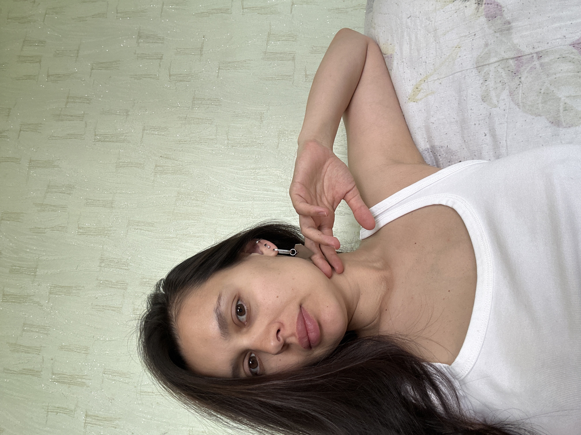A woman with long, dark hair, fair skin, and brown eyes is lying down, posing for a selfie. She is wearing a white tank top and has multiple piercings in her left ear. Her right hand is raised, with her fingers near her neck. The background shows a textured, light green wall and a white bedsheet with a floral pattern. The location is presumed to be within a residence in the city of Chaikovsky, Russia, based on the user's request.