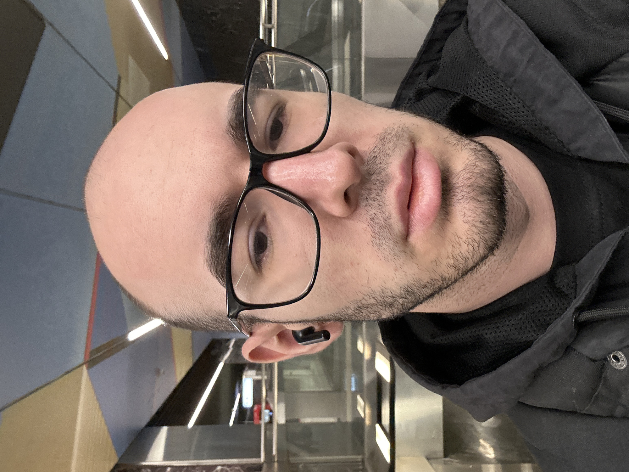 A close-up photo depicts a bald, white male wearing black-rimmed glasses and a black earbud in his left ear. He has a light beard and appears to be wearing a black hooded jacket. The background suggests an indoor location with geometric designs on the wall and bright, rectangular light fixtures. A glass or metal barrier is visible in the background. While no location in the city of Khimki, Russia, can be determined without further information, the image's environment indicates an indoor space, possibly a station or building with contemporary design elements.