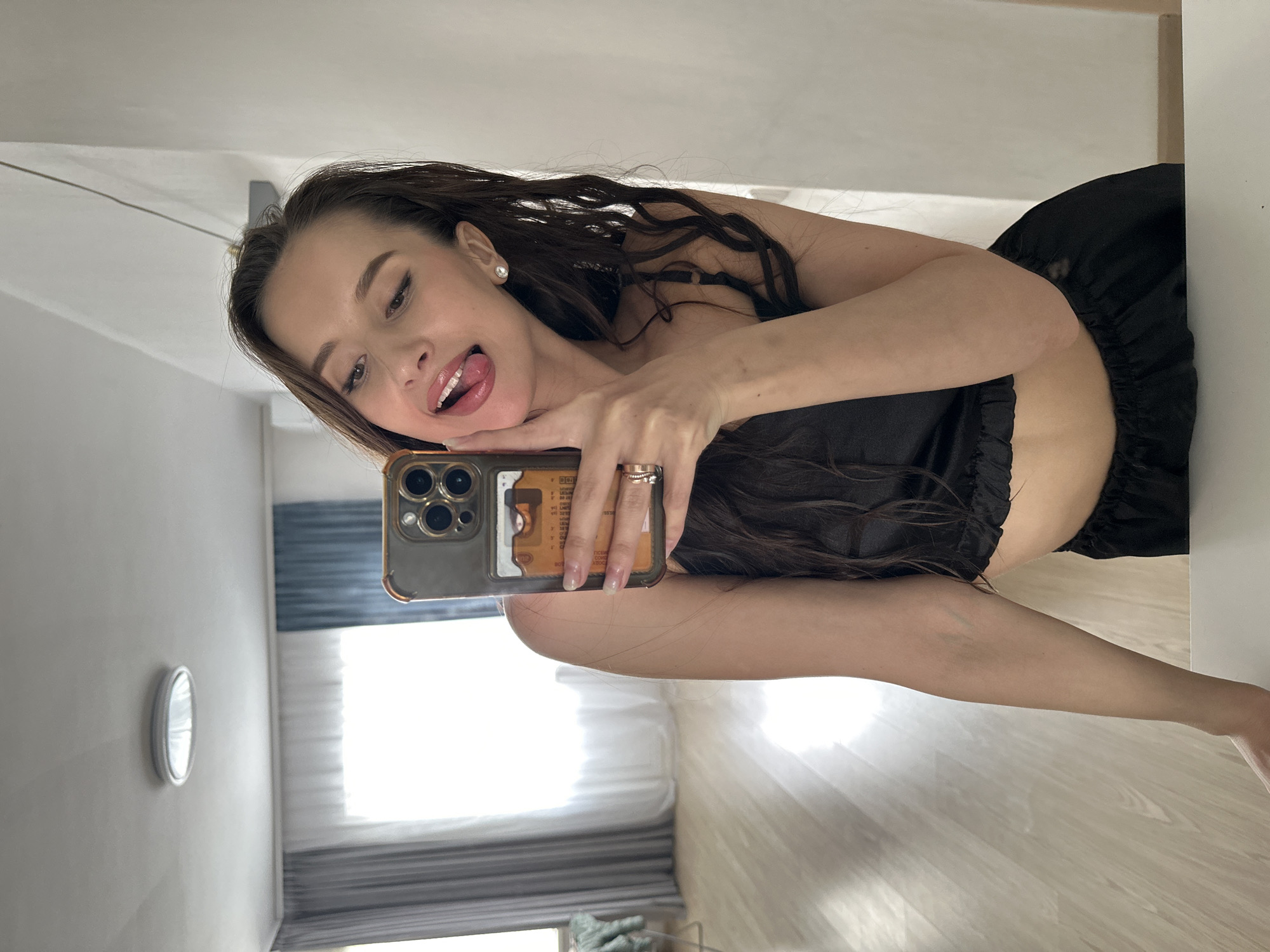 A young woman is taking a selfie in what appears to be a bedroom or living space. She's wearing a dark, possibly black, top and dark bottoms. She has long brown hair, and her tongue is sticking out. She's holding a cell phone, possibly an iPhone, with a visible case. The background includes a window with sheer white curtains and a partial view of the outside. A round object is attached to the wall and looks like a clock. The floor is light wood. It is not possible to determine the location in the city of Yekaterinburg, Russia, from the image.