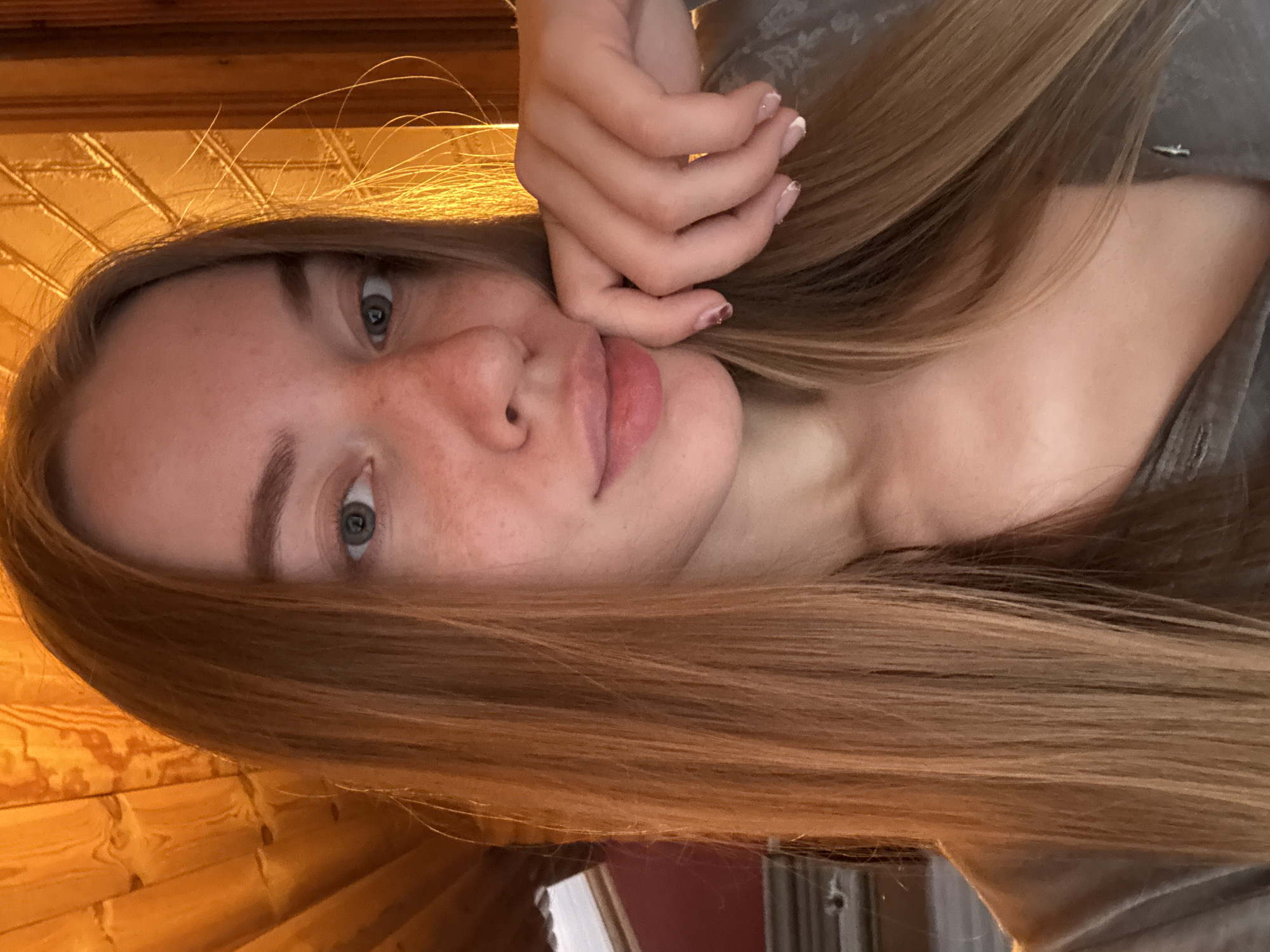 A young Caucasian woman with long, straight, light brown hair is shown in a close-up selfie. She has fair skin with freckles, blue eyes, and is resting her right hand on her chin. She is wearing a gray shirt. The background appears to be an interior with wood paneling, illuminated by warm, artificial light, creating a cozy atmosphere. There are no discernible urban elements or specific locations within the frame.