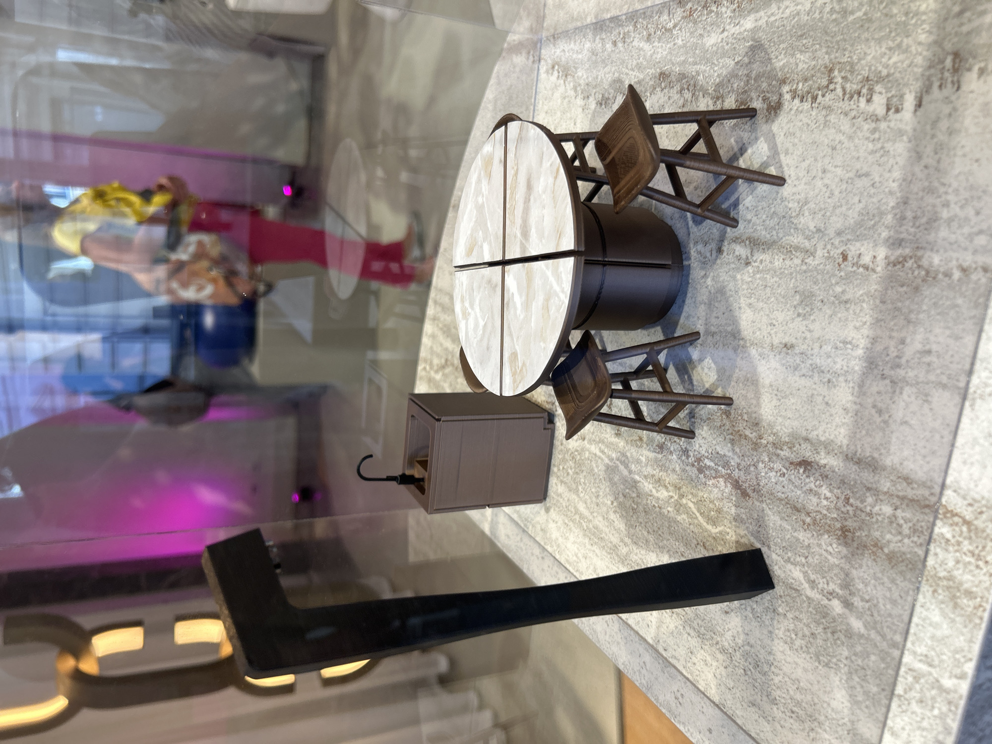 The image displays a miniature or model scene, viewed through a reflective glass surface. The central focus is a small, circular table with a light, marble-patterned top divided into four sections, accompanied by two dark brown chairs with a simple, wooden-like design. Next to the table is a small, rectangular brown box with a black hook protruding from it, possibly a miniature lantern or decorative item. The miniature set rests on a textured, light-grey and beige surface resembling stone or concrete.
In the foreground, a large, dark, angular object, likely part of a display stand, extends upwards from the bottom left. The reflective glass surface prominently features reflections of an indoor environment. These reflections include distorted figures of people, with one person's upper body visible in yellow and another in red, suggesting observers of the display. Vibrant artificial lighting is also reflected, with distinct pink/magenta glows and warm, golden-yellow light that appears to form parts of illuminated letters or a sign, though no full text is discernible (possible fragments like "CL").
The scene depicts a static, decorative exhibit, possibly a diorama or model display, situated in an indoor, likely public or commercial setting such as a gallery or retail space. The activity is one of observation. There are no direct indications of time of day or weather. Given the provided context, the location is Milan, Italy.