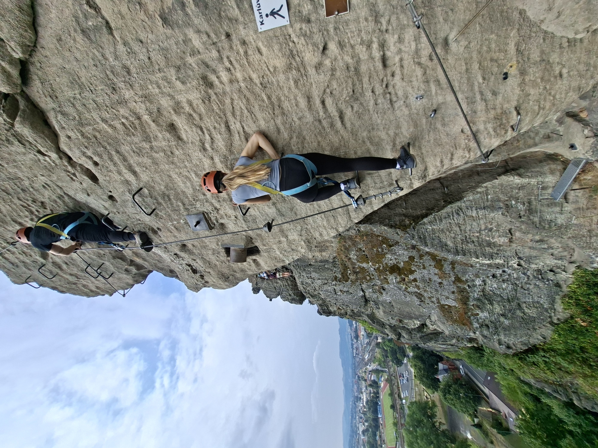 The image depicts a rocky cliff face with two individuals engaged in climbing, likely on a via ferrata route. The person in the foreground, a woman with blonde hair wearing a grey long-sleeved shirt, black leggings, a blue climbing harness, and an orange helmet, is ascending the rock face using metal rungs and cables. She is positioned mid-climb, with her right leg on a rung and her left hand gripping a cable. Above her, further up the cliff, a second person, a man wearing a black t-shirt, black pants, a yellow and blue climbing harness, and an orange helmet, is also climbing, his back to the camera.
The rock face itself is rough and textured, with various ledges and crevices. Metal fixtures, including rungs and cables, are bolted into the rock to facilitate climbing. A sign with a walking figure and text, partially obscured but potentially reading "Karlův," is visible on the right side of the rock face.
In the background, a panoramic view of the city of Děčín, Czechia, unfolds. The cityscape includes buildings, a railway track with a train, roads with cars, a stadium with a track, and a body of water. The sky is overcast with scattered clouds. The vantage point suggests the climbers are high above the city, offering a dramatic perspective. The overall scene captures an outdoor adventure activity set against an urban backdrop.