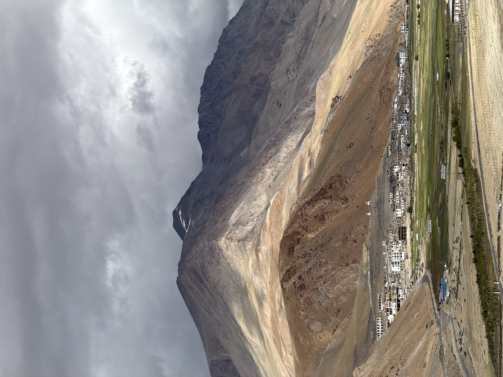 The image depicts the remote Himalayan village of Karzok, India, nestled in a valley at the base of towering, arid mountains under a dramatic, cloudy sky. The village is characterized by its cluster of white-washed buildings, many with flat roofs, interspersed with green agricultural fields. A winding road leads into the village, with several vehicles visible on it and near a small cluster of buildings. The landscape is stark and rugged, with the mountains exhibiting layers of tan and reddish-brown rock, partially illuminated by shafts of sunlight breaking through the clouds. Two small white stupas are visible on the higher slopes behind the village, common in Buddhist regions of the Himalayas. The overall impression is one of isolation and resilience in a harsh, yet beautiful, natural environment.