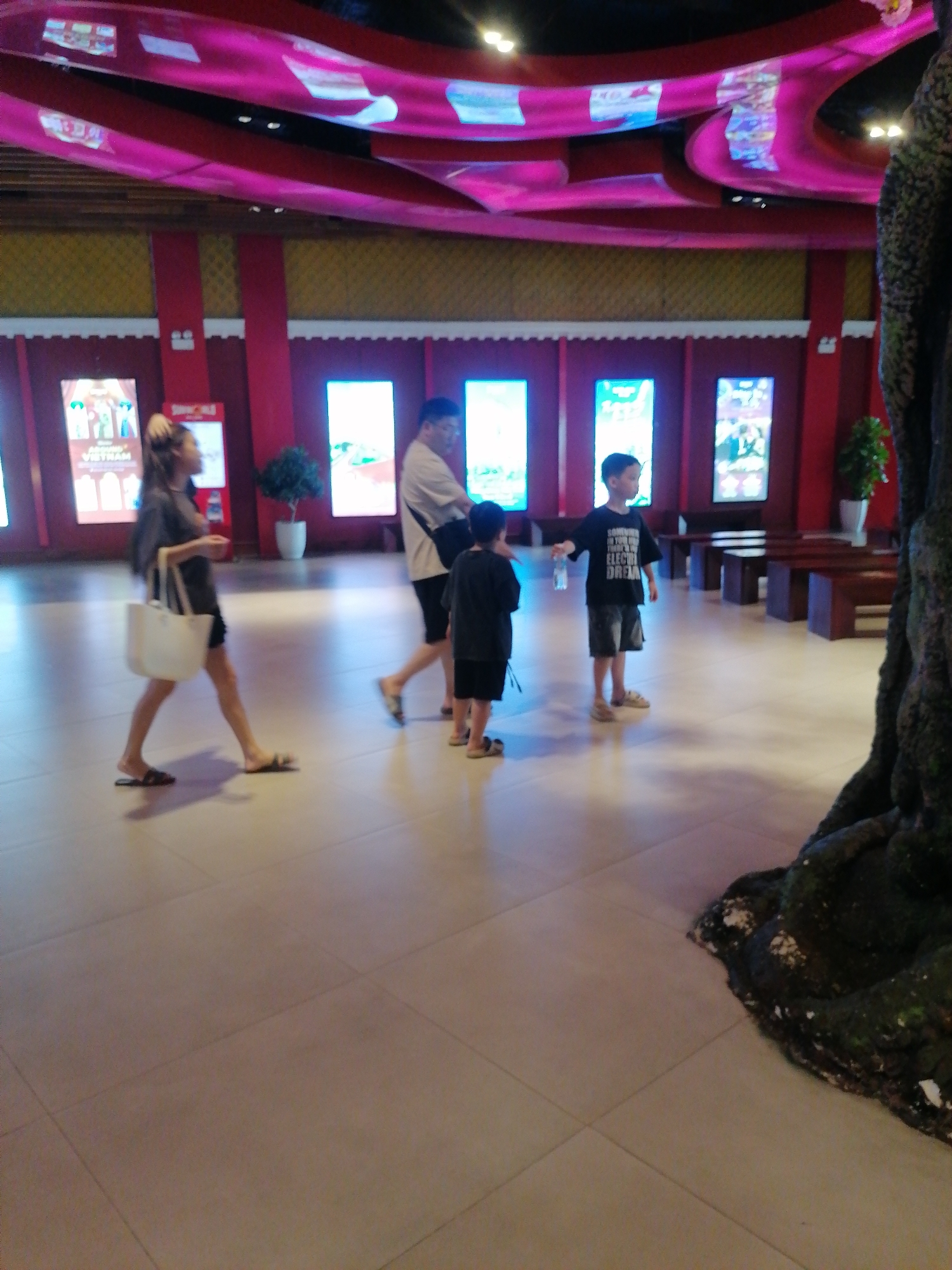The image shows a brightly lit indoor space in Hong Gai Ward, Vietnam. Visible are tiled floors, red pillars, and decorative ceiling elements that appear to be video displays with colorful abstract content. Several people are present: a woman is walking towards the left, carrying a white bag. A man and two boys are standing near each other, also toward the left of the frame. Benches and potted plants are situated along the back wall. The background also features video displays with content that is not easily discernible. In the foreground to the right is a large, textured artificial tree trunk.