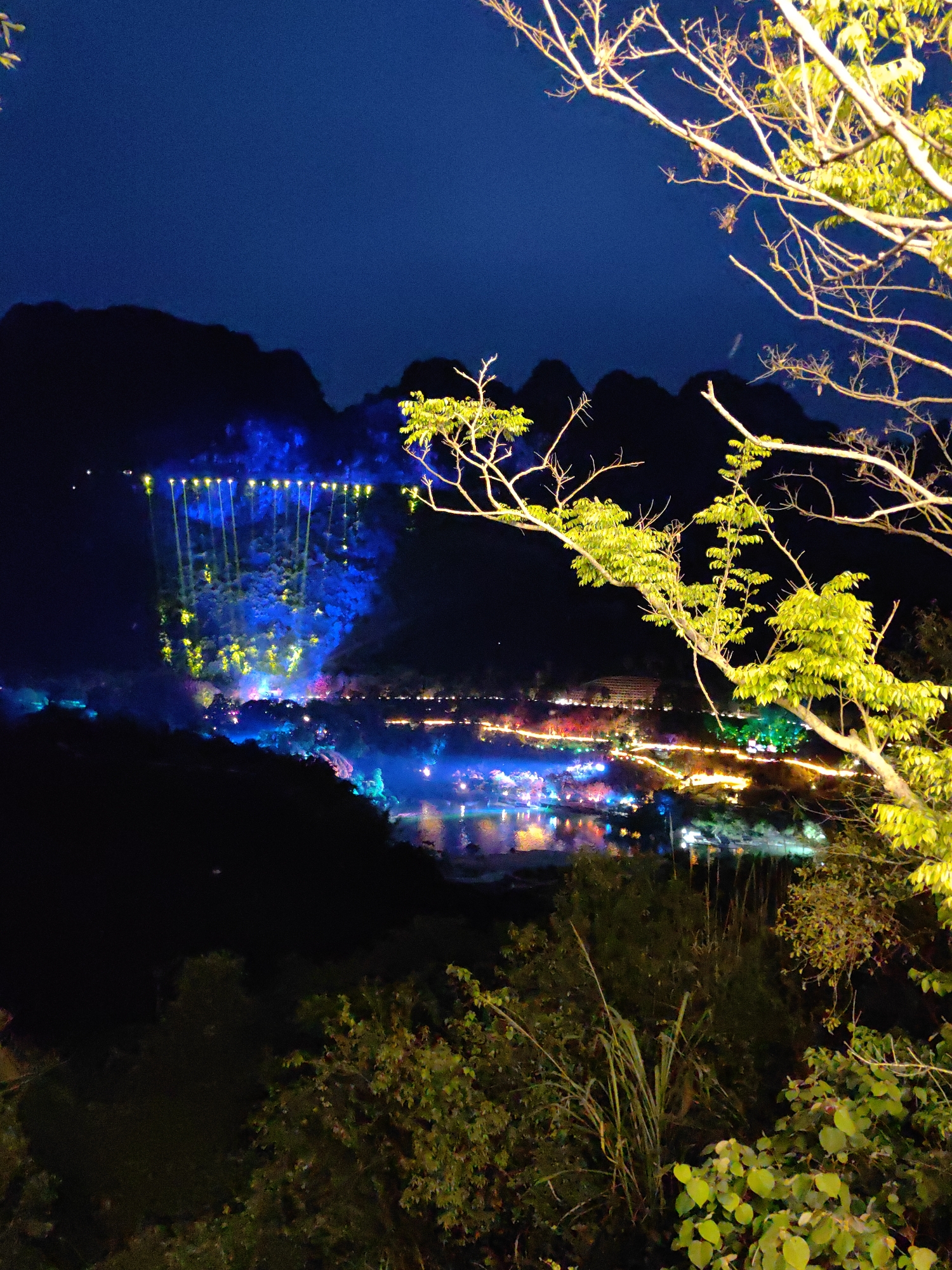 Nighttime view of Xã Đàm Thủy, Vietnam, featuring illuminated mountains, a body of water reflecting colored lights, and trees in the foreground. The mountains in the background are dark, with blue and yellow spotlights shining on them. The water reflects blue, pink, and green lights from the buildings and structures along the shore. In the foreground, trees with green leaves and bare branches are visible, slightly illuminated.